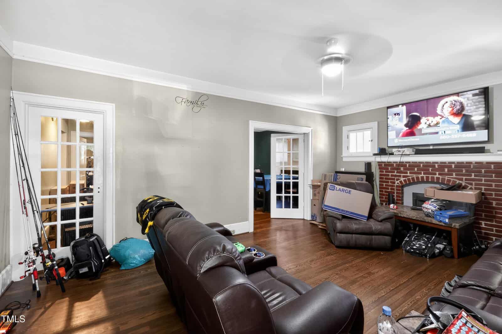 A living room with brown leather couches, a TV above a brick fireplace, fishing rods, boxes, and bags scattered around. The walls are light-colored with a "Family" decal, and there are two French doors leading to adjoining rooms.