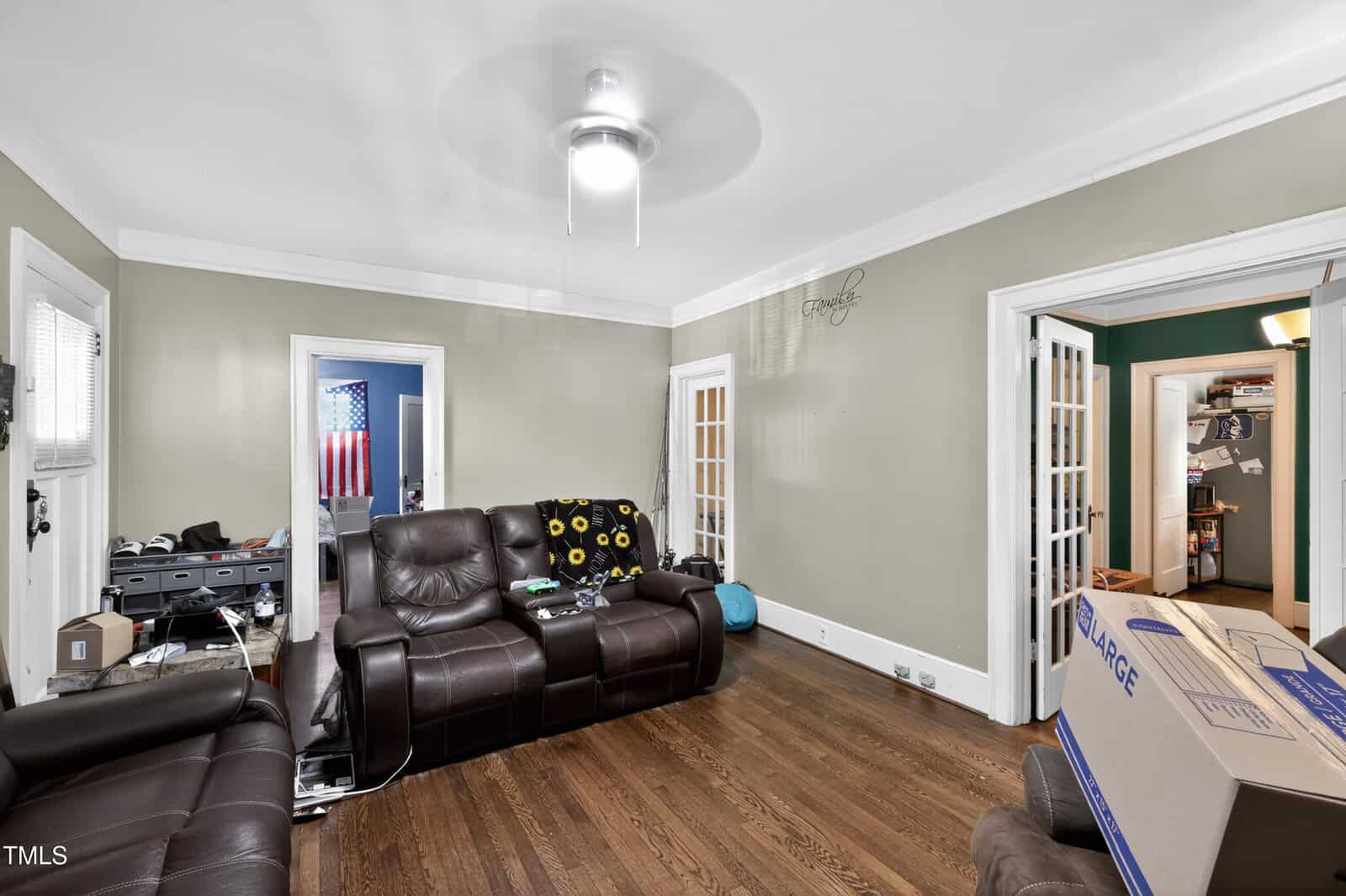 A living room with hardwood floors, brown leather sofas, moving boxes, and French doors leading to other rooms. A ceiling fan with a light is centered on the white ceiling. The walls are painted beige.