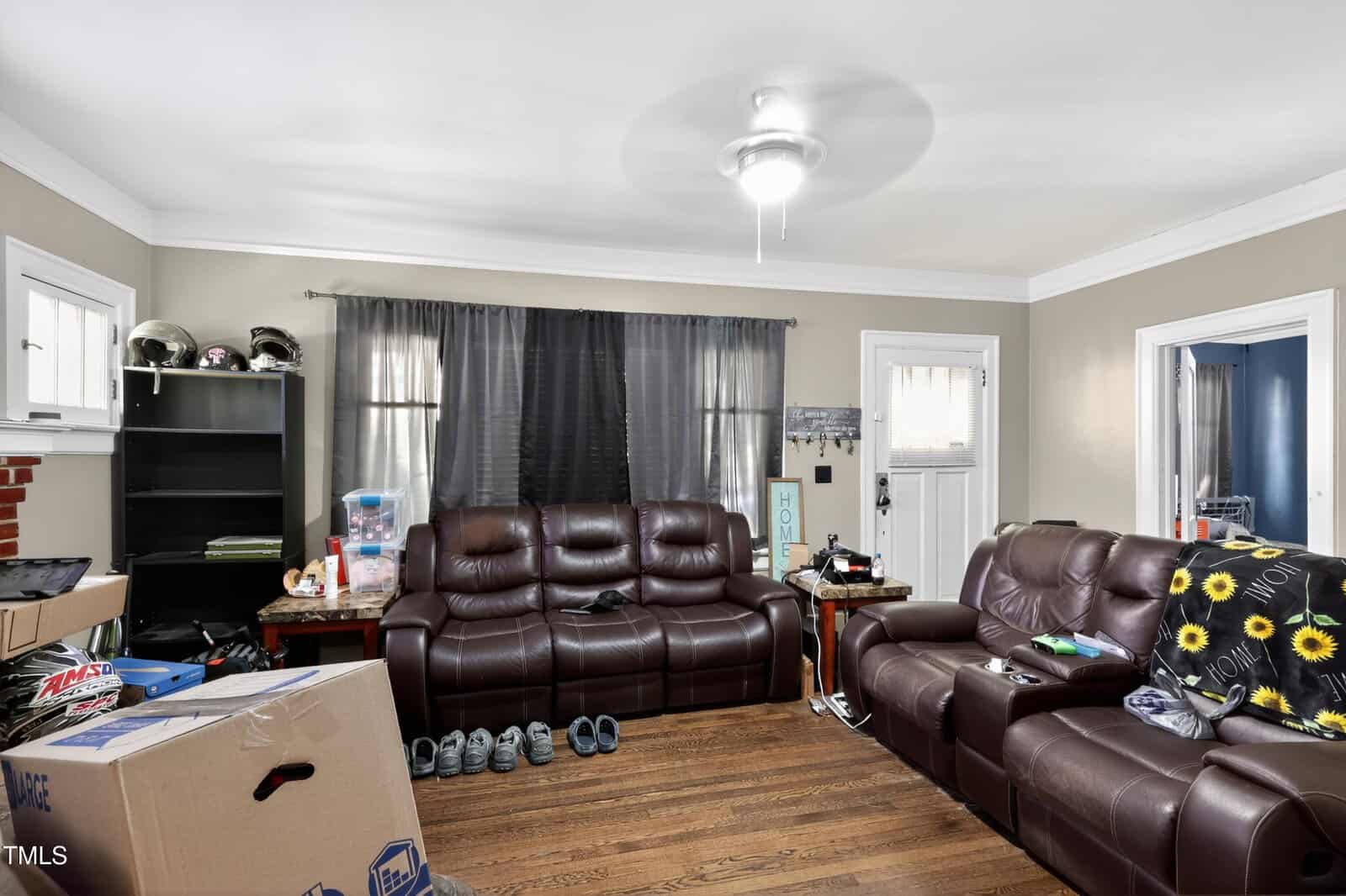 A living room with brown leather sofas, a wooden floor, and a ceiling fan. There are unpacked boxes, shoes, and various items scattered around, with shelves and windows in the background.