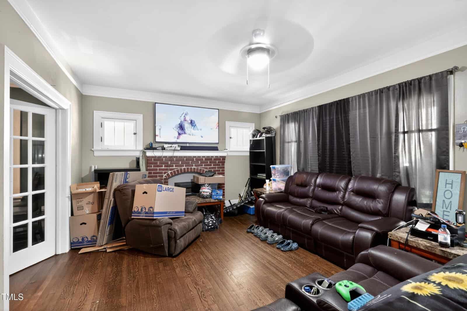 A living room with brown leather sofas, a brick fireplace, moving boxes, a TV mounted above the fireplace, dark curtains, and scattered personal items on the floor and tables.