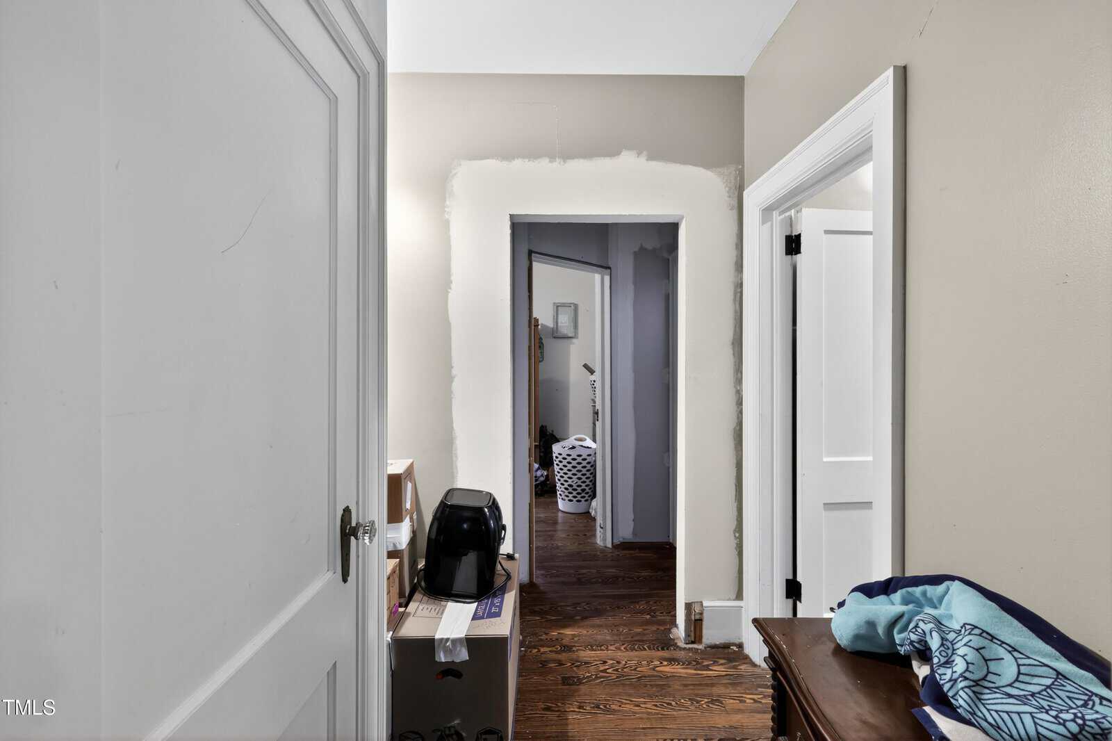 A hallway with unfinished drywall around a doorway, hardwood floors, a laundry basket in the next room, and various items including a small appliance and boxes along the walls.