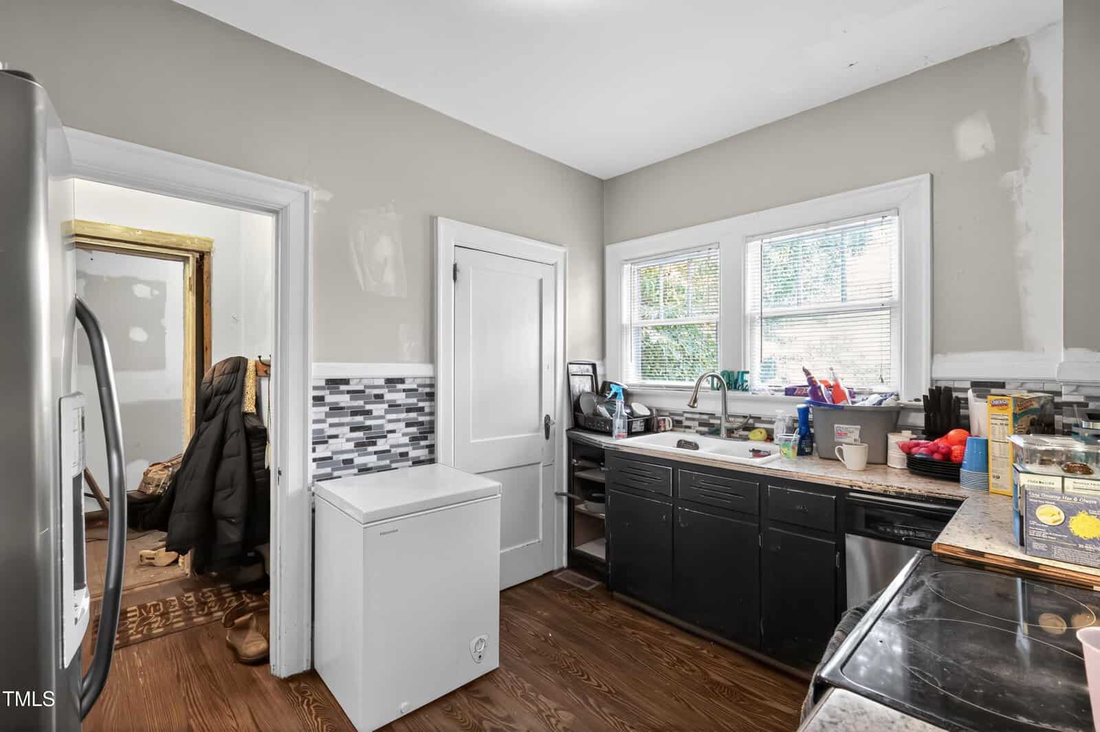 A kitchen with dark lower cabinets, a small white chest freezer, a tile backsplash, and a window above the sink. The countertop holds dishes, fruit, and various groceries. A coat and bags hang in the adjacent room.