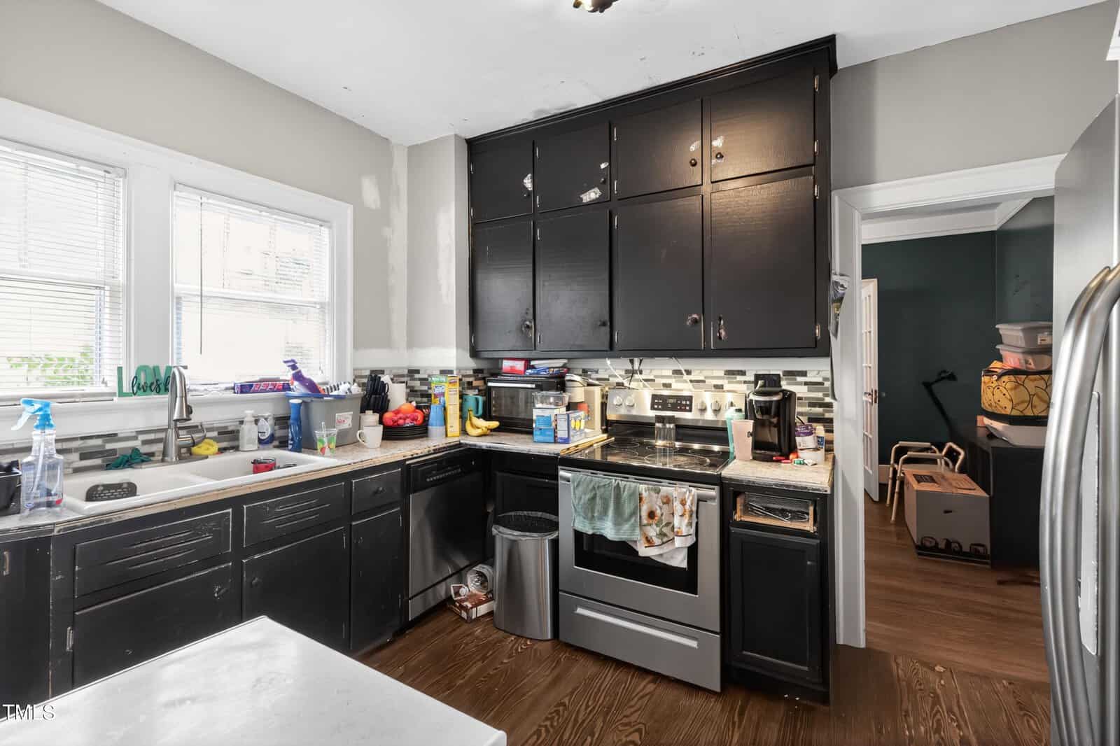 A kitchen with black cabinets, stainless steel appliances, a cluttered countertop with food and cleaning supplies, and a partially open door leading to another room with moving boxes.