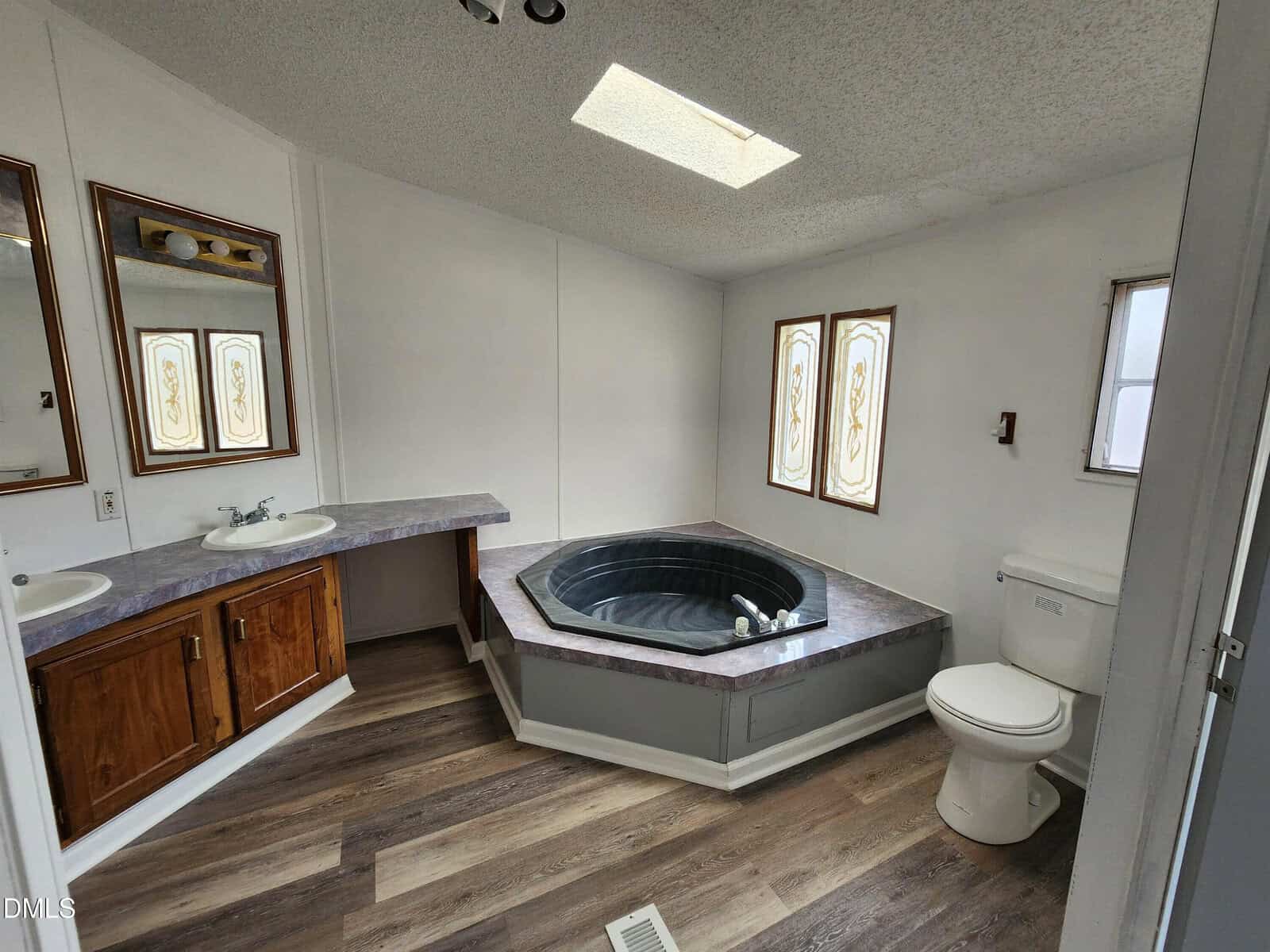 A bathroom with wood-look flooring, a built-in corner soaking tub, a double sink vanity with wood cabinets, a toilet, mirrors, and stained glass windows. A skylight is above the tub.