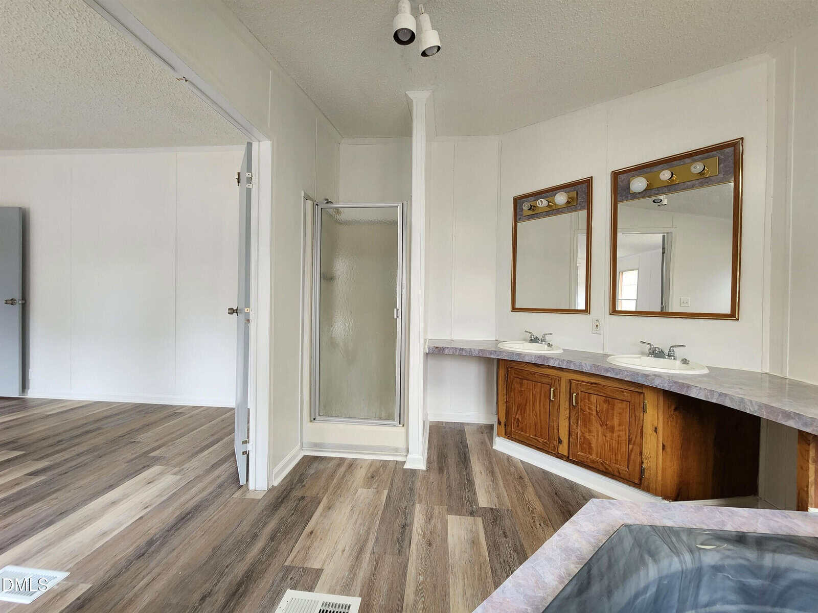 A bathroom with wood-style flooring, a corner shower with a glass door, two sinks with mirrors above a wooden cabinet, and part of a bathtub visible in the lower right corner.