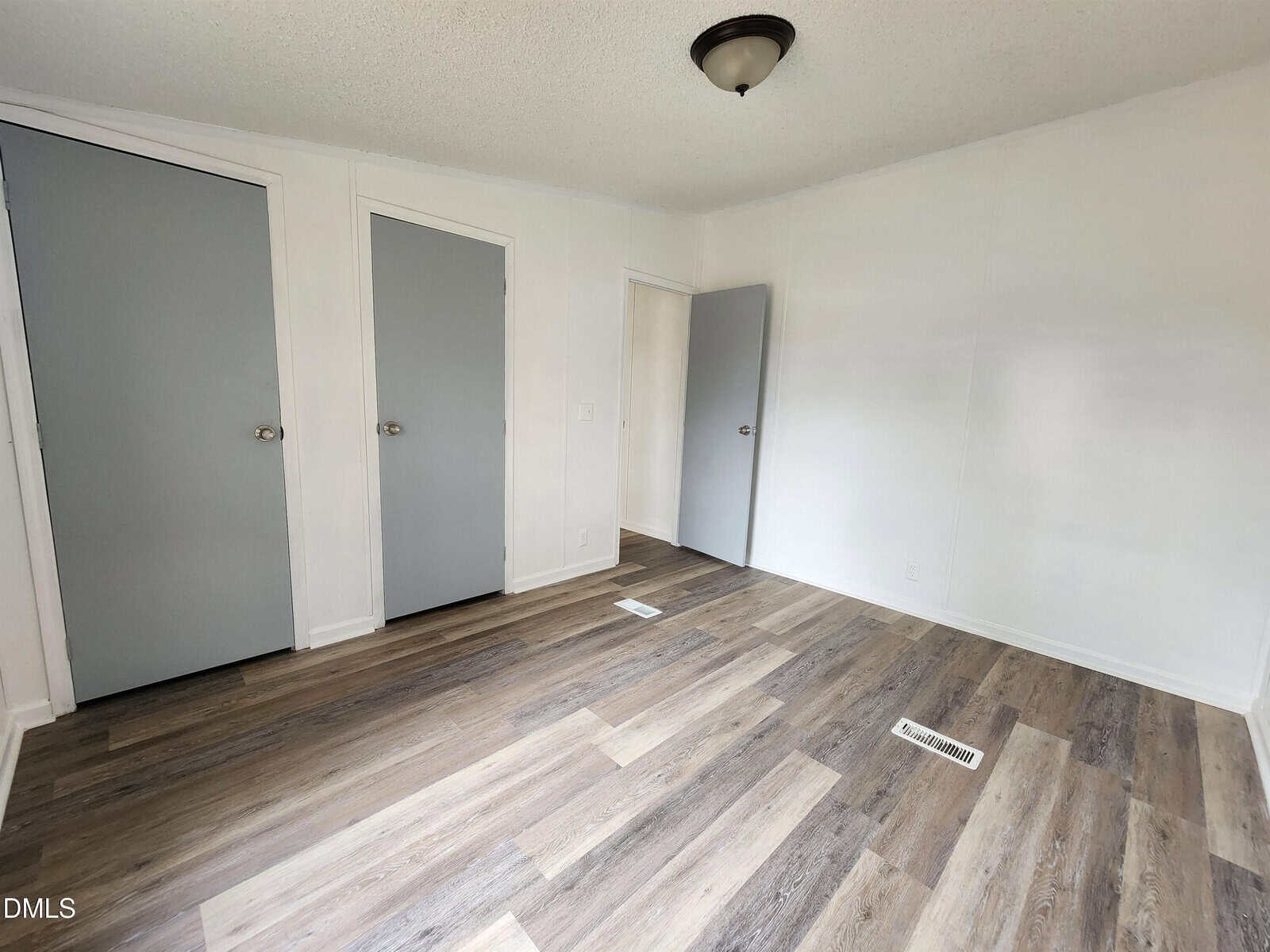 Empty room with white walls, a light fixture on the ceiling, two closed gray closet doors, one open gray door, and wood-patterned vinyl flooring with a floor vent.