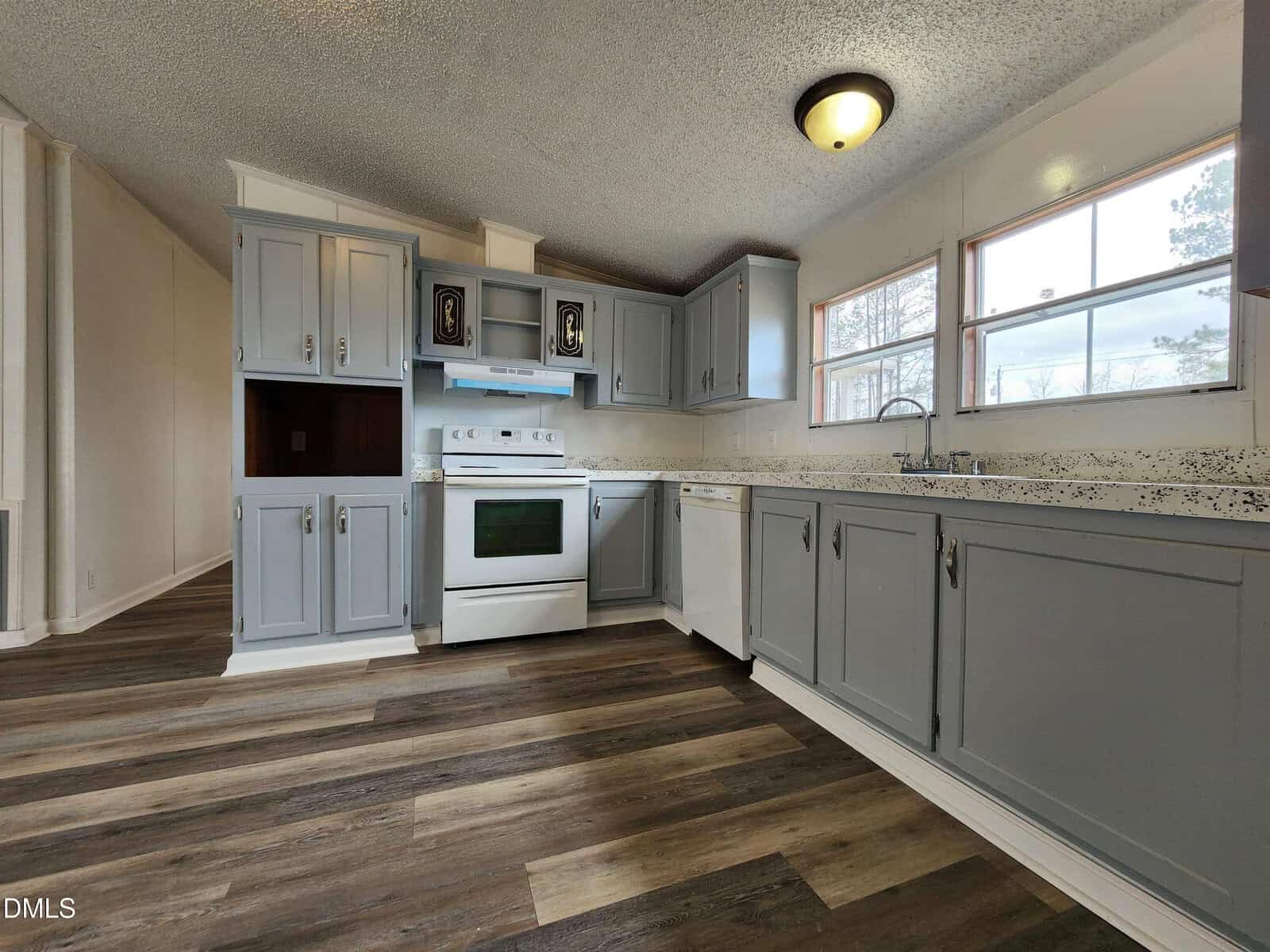 A kitchen with gray cabinets, white appliances, wood-look flooring, a speckled countertop, and large windows letting in natural light. The stove and oven are centered between cabinetry.