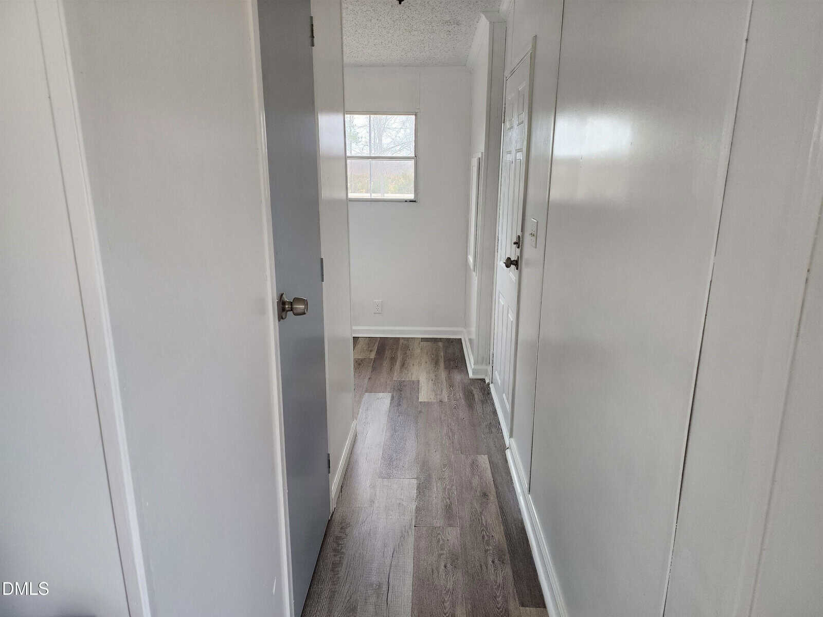 A narrow hallway with white walls and two closed doors, featuring dark wood-look flooring and a window at the far end letting in natural light.