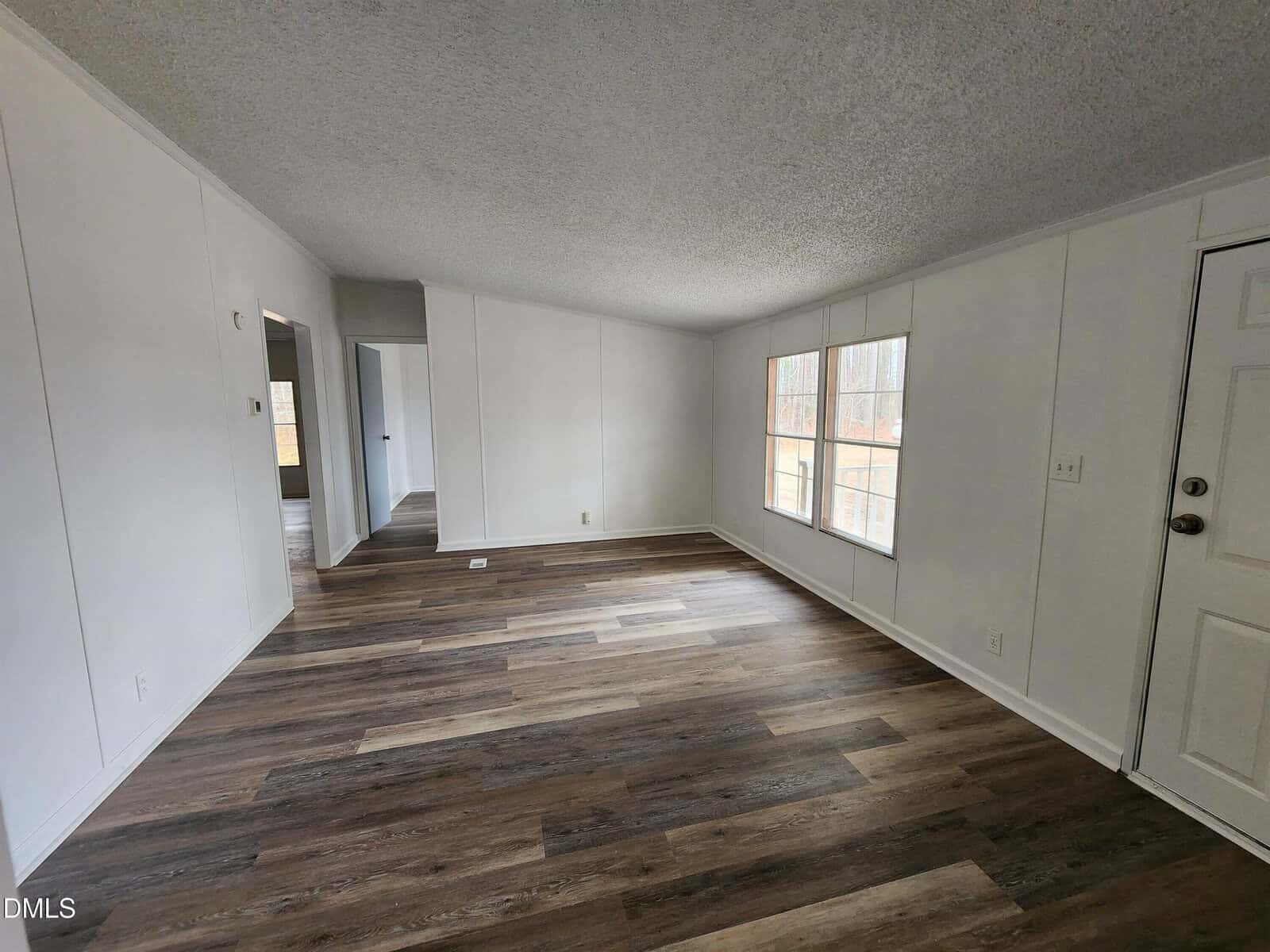 Empty room with white walls, wood-style flooring, two large windows, and a closed white door. A hallway and another room are visible in the background. The space is unfurnished and well-lit by natural light.