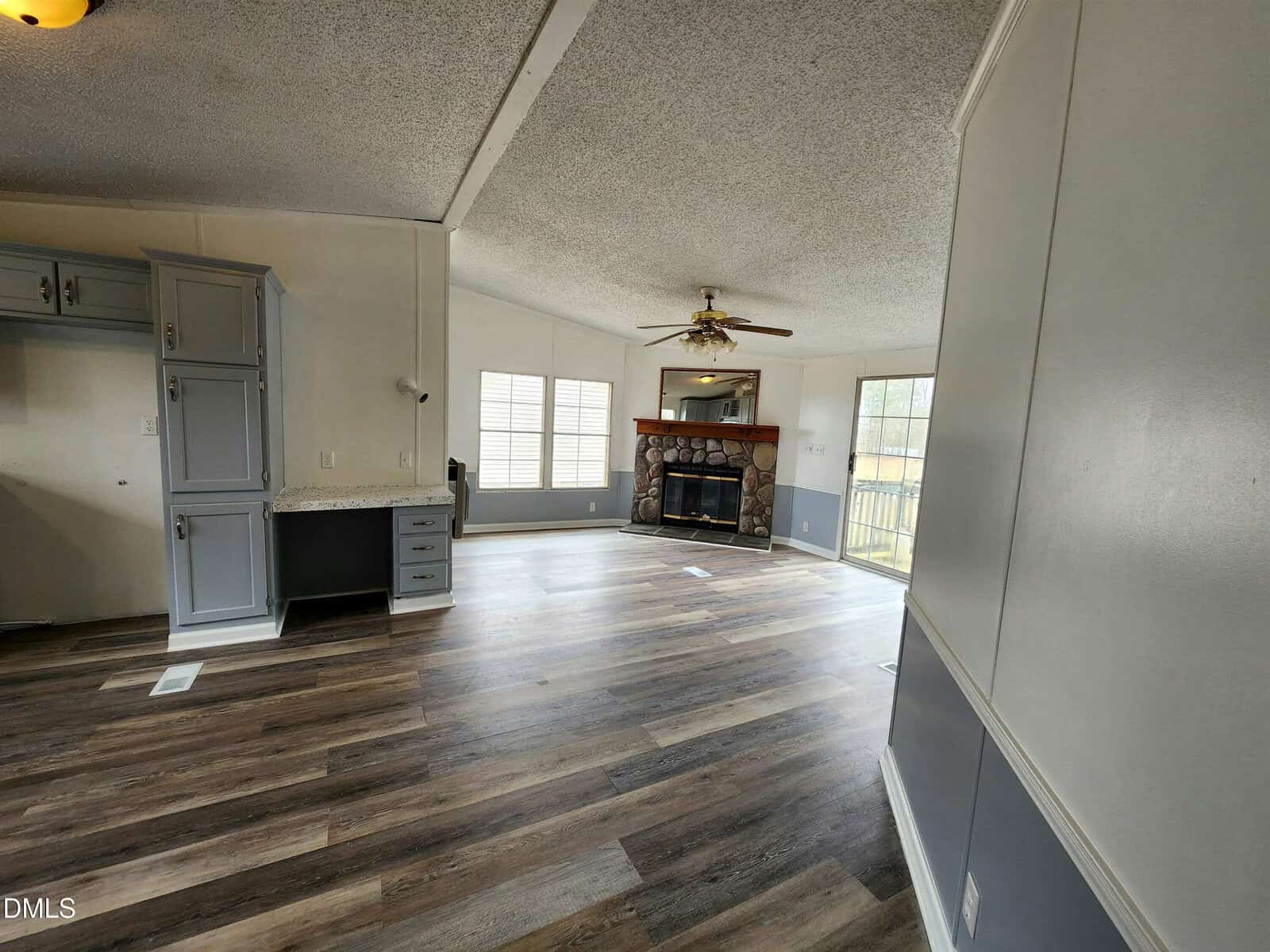 A spacious living area with wood-look vinyl flooring, grey built-in cabinets, a ceiling fan, large windows, and a stone fireplace against the far wall. Natural light fills the room through the windows and glass door.