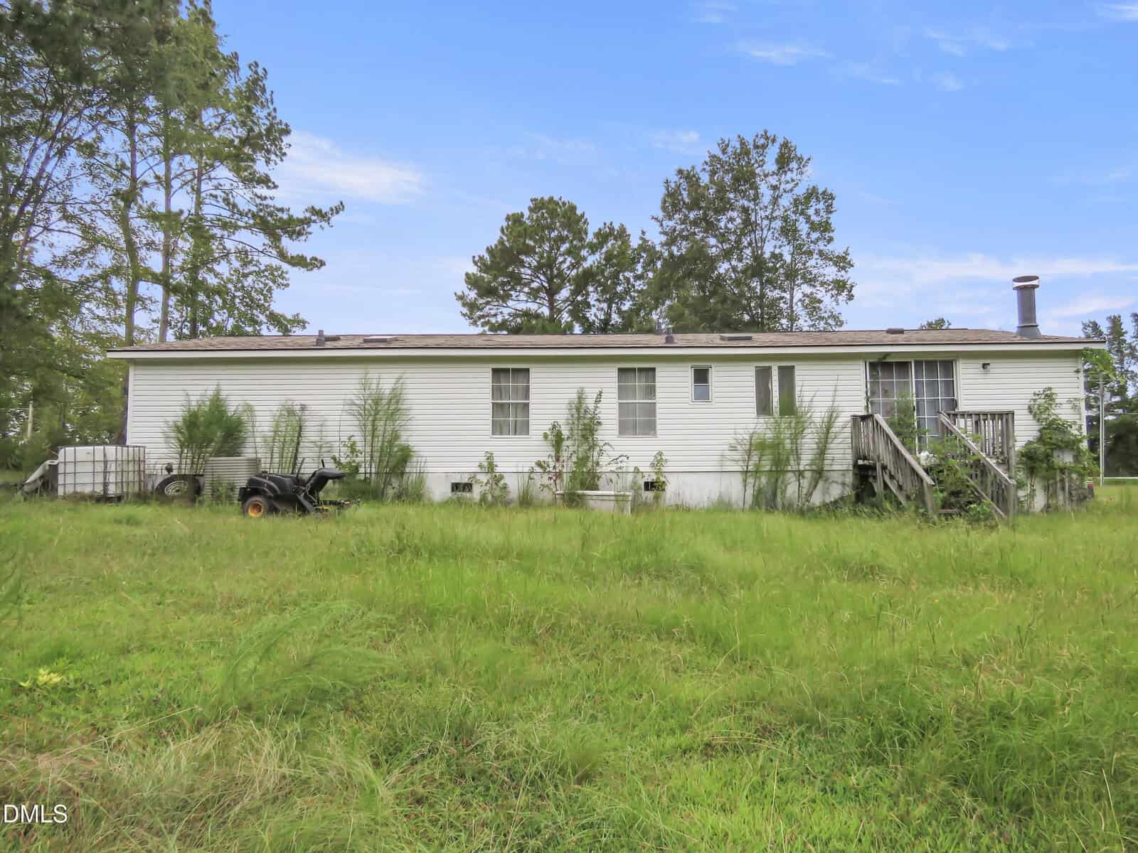 A white manufactured home with overgrown grass and bushes in the yard, a small trailer and cart near the house, steps leading to a door, and trees in the background under a blue sky.