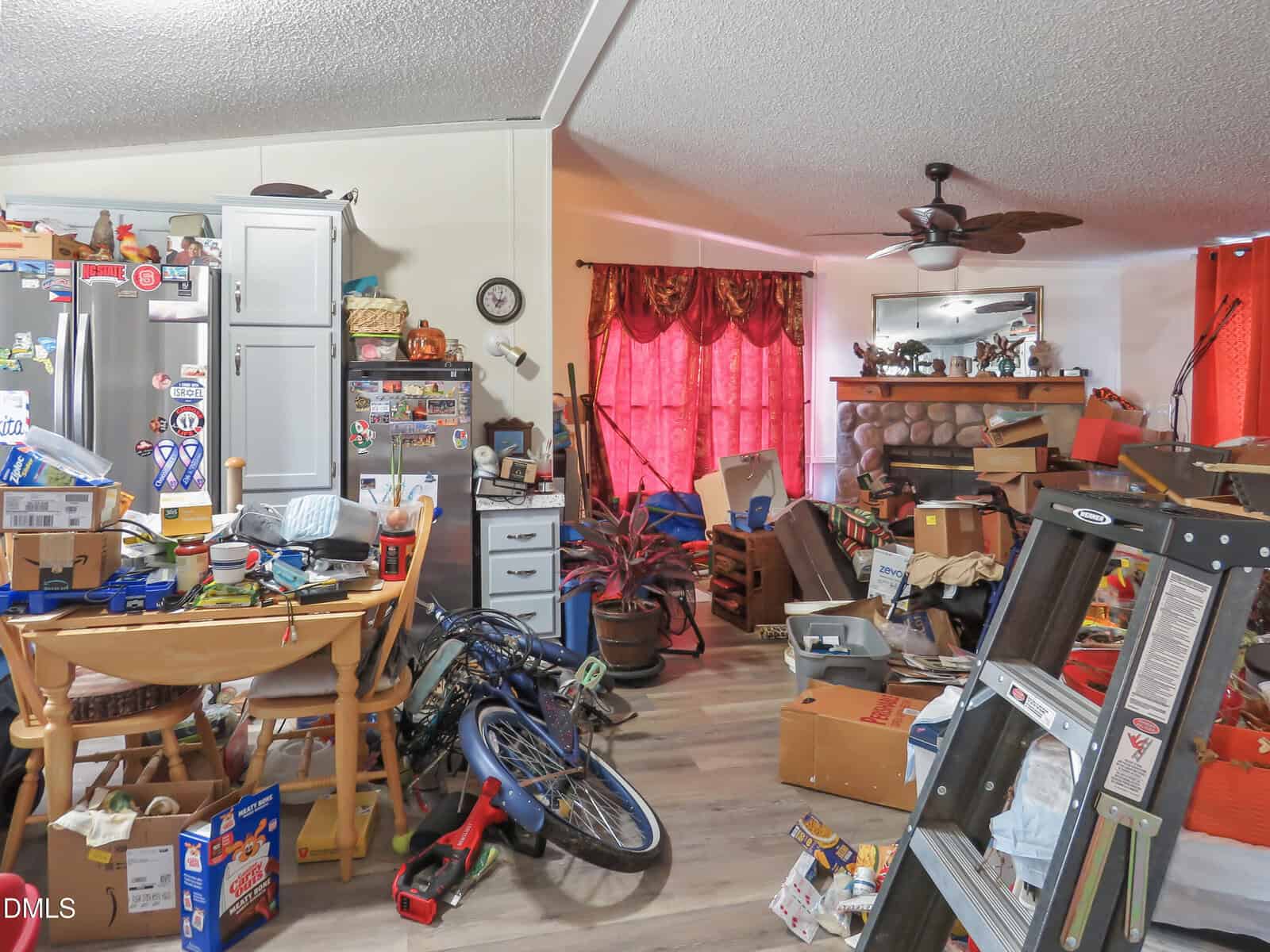 A cluttered room with various items scattered, including boxes, a bicycle, a ladder, kitchen furniture, and appliances. Red curtains cover windows, and shelves are filled with household goods and decorations.