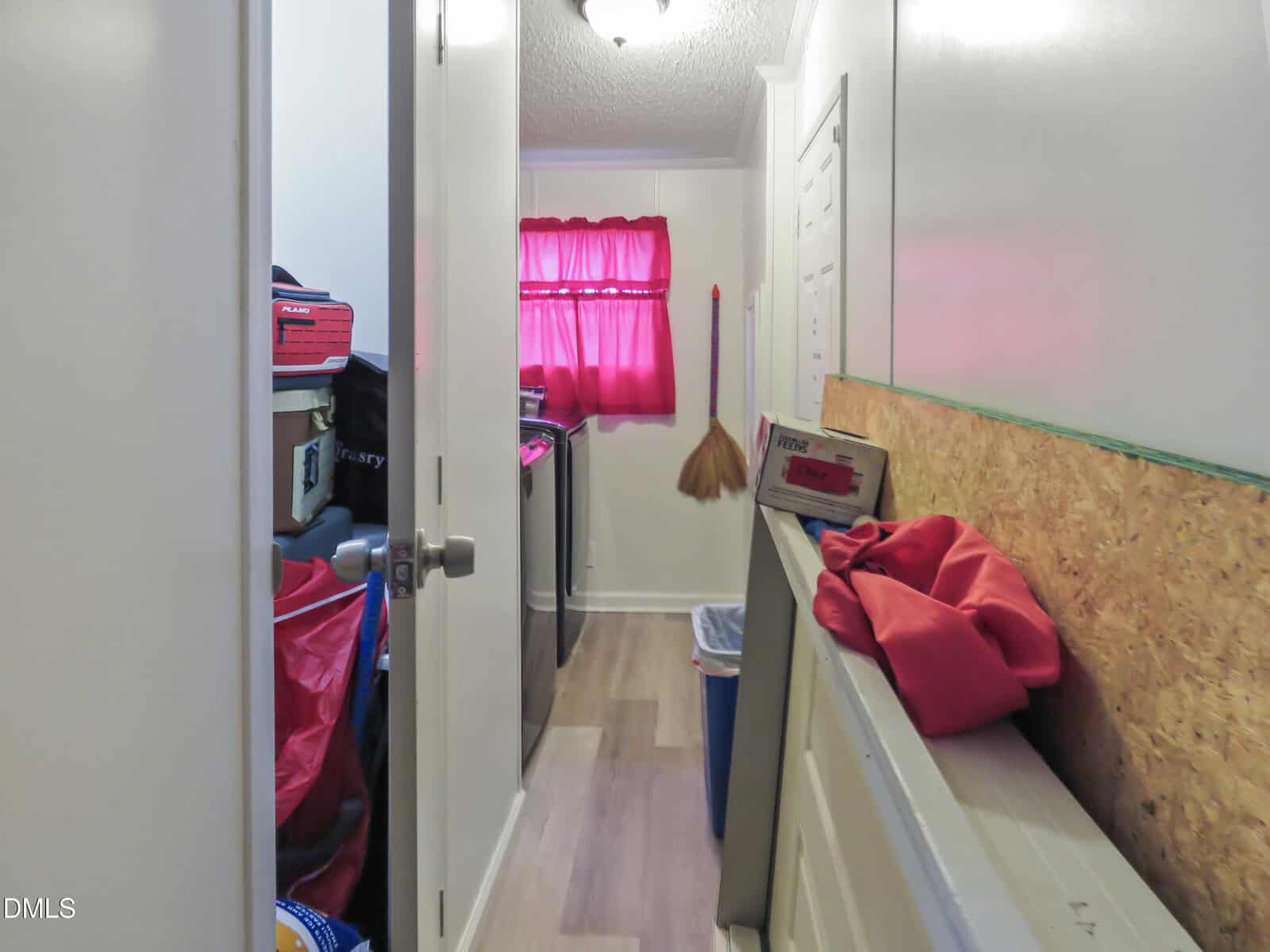 A small laundry room with a washer and dryer, cleaning supplies, storage bins, a broom hanging on the wall, and a window with a red curtain. A countertop holds a box and a red cloth.
