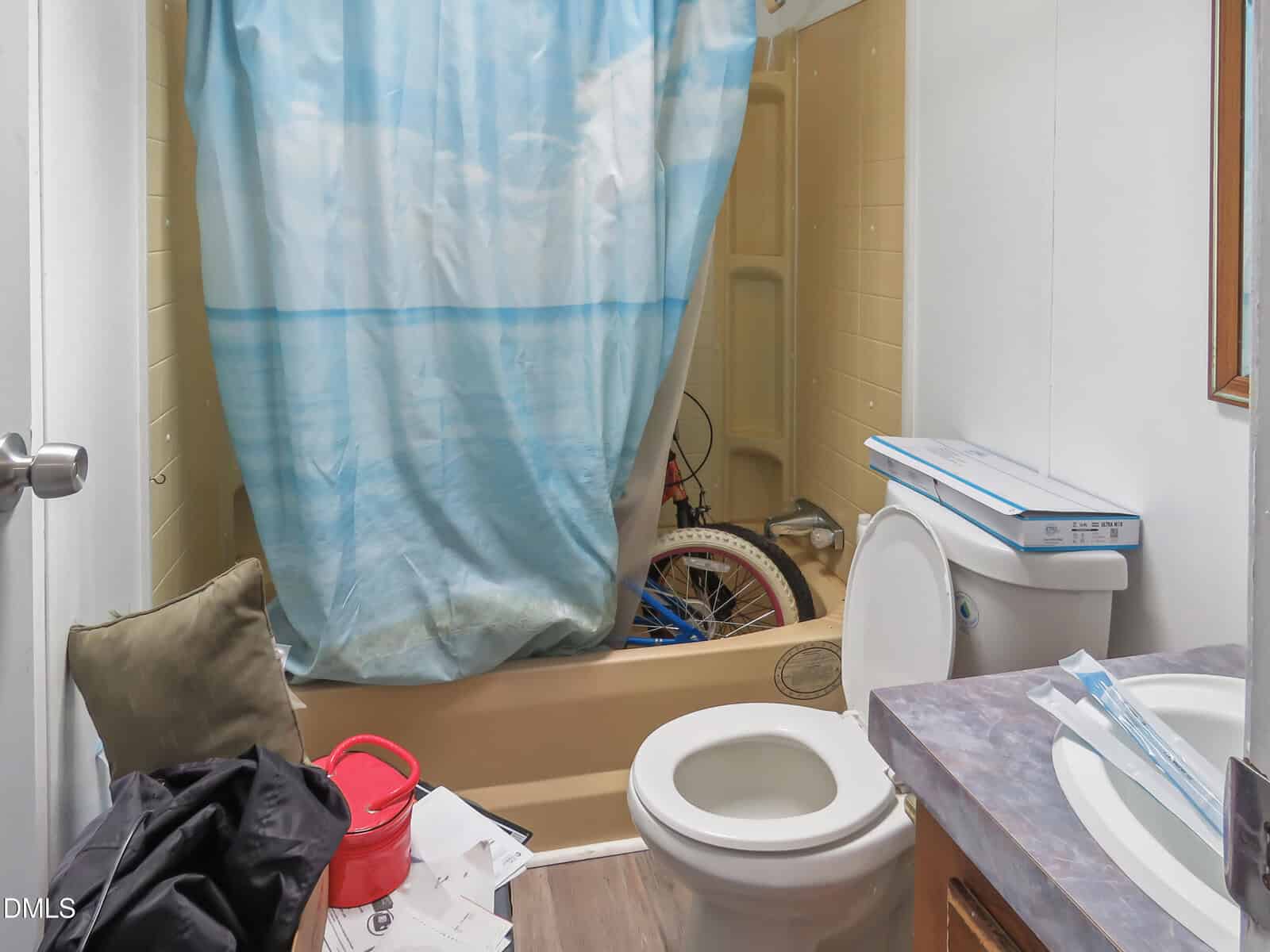 Small bathroom with tan tub, blue shower curtain, white toilet, cluttered counter, and a bicycle partially visible in the bathtub. Various items and papers are scattered around.