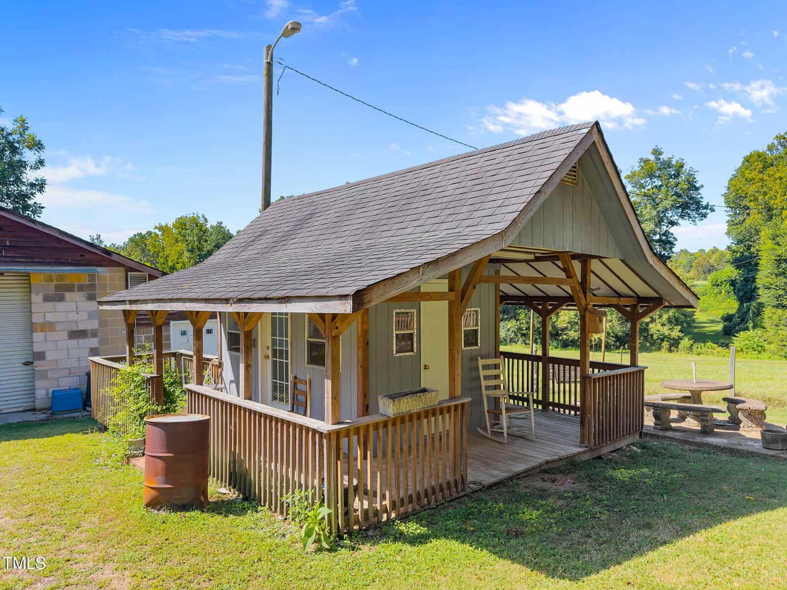 A small wooden cabin with a covered porch and railing sits on a grassy yard. The porch has a rocking chair and a table. Nearby are a metal drum, picnic table, and trees under a clear blue sky.