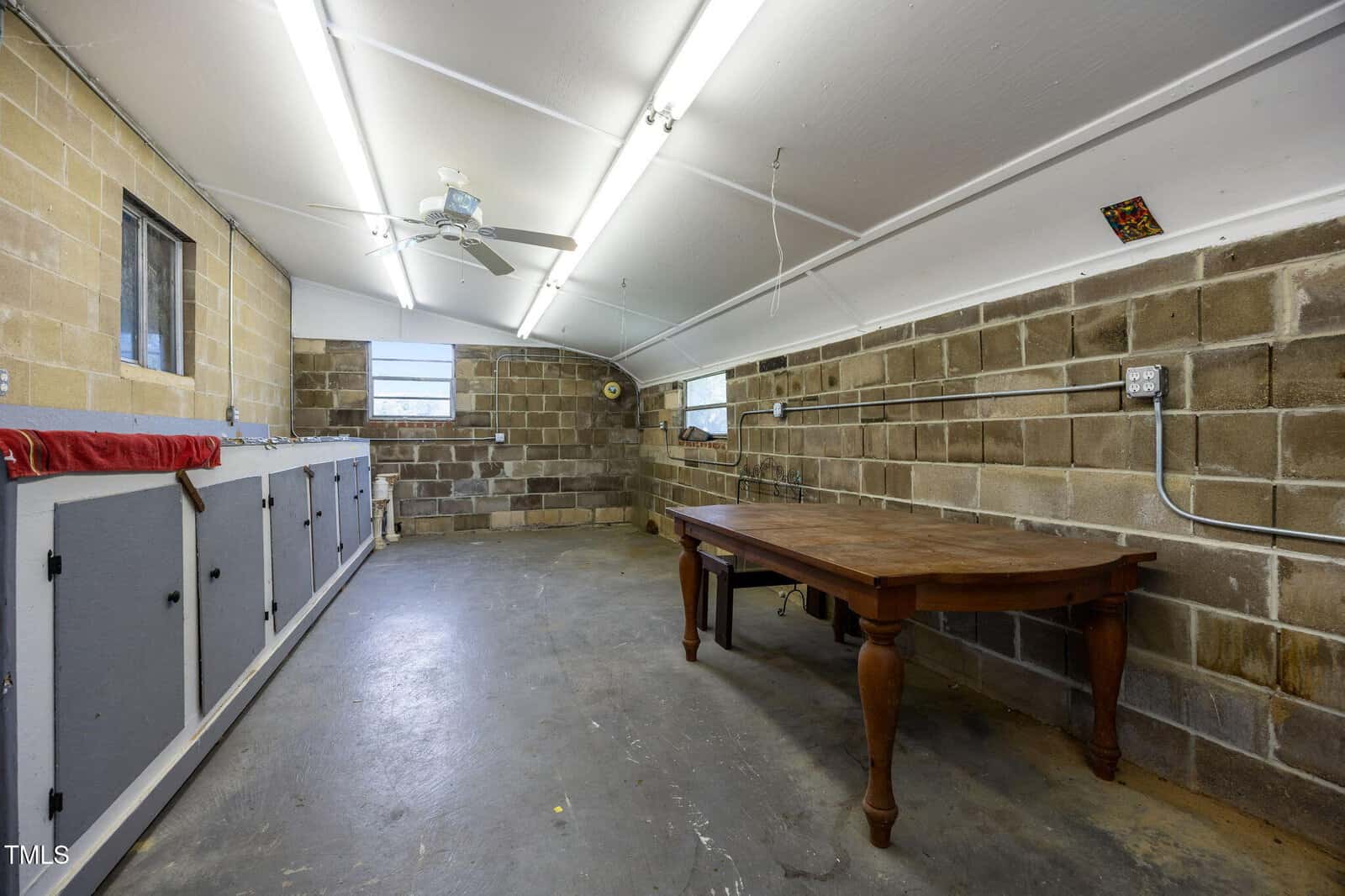 A long, narrow room with cinder block walls, fluorescent ceiling lights, two small windows, a ceiling fan, a row of cabinets on the left, and a wooden table on the right. Electrical wiring is visible on the walls.
