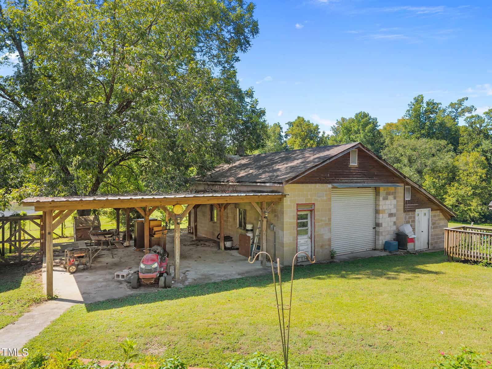 A single-story building with a metal garage door and covered carport area, containing a red lawn mower and various equipment, sits on a grassy yard with trees in the background under a clear sky.
