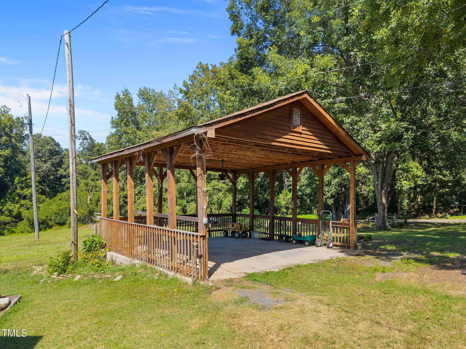 A wooden open-air pavilion with a pitched roof and railing stands on a grassy area surrounded by trees. There is playground equipment stored inside and power lines are visible nearby under a blue sky.