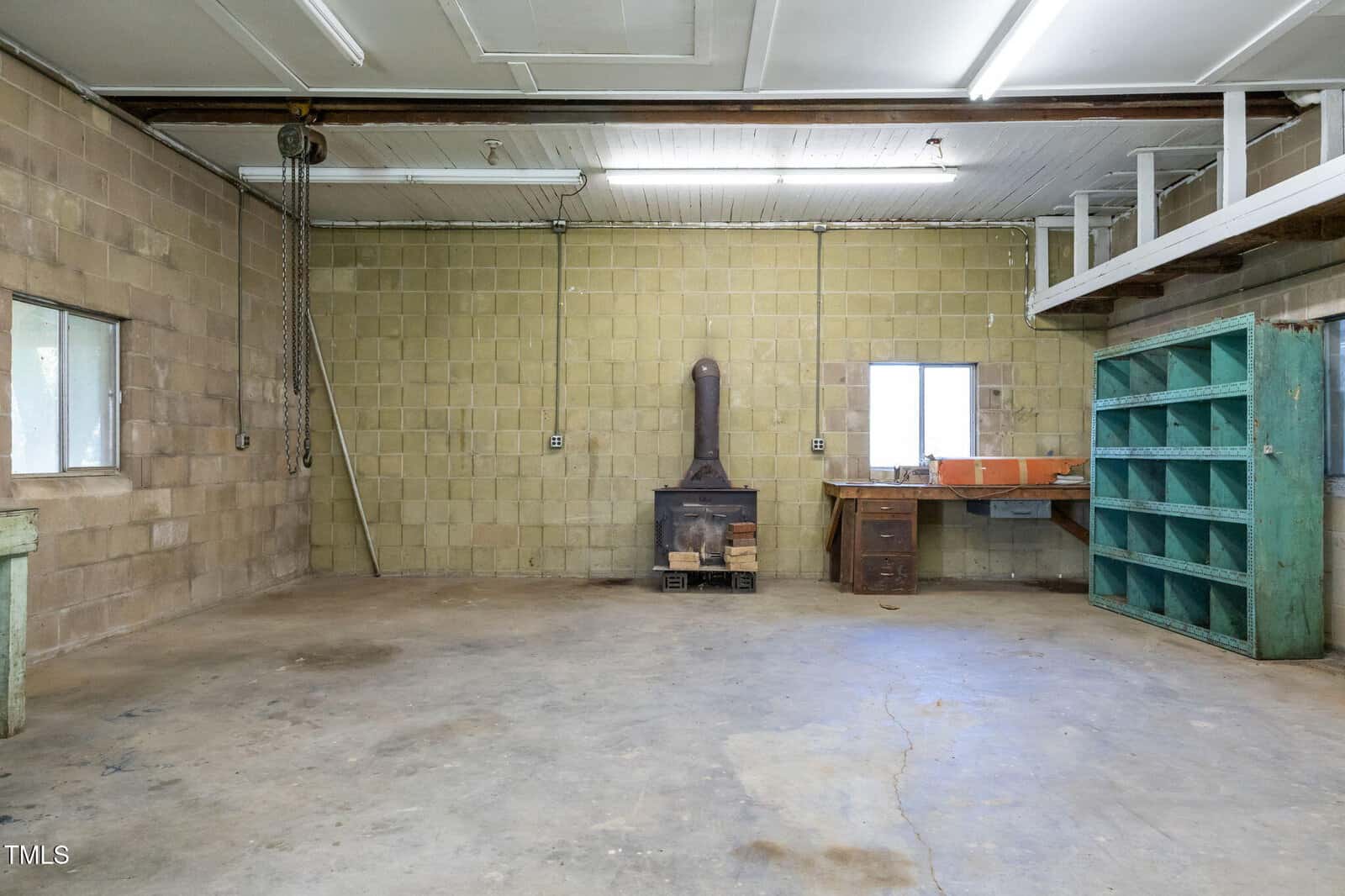 A spacious workshop with concrete floors, yellow cinder block walls, a wood stove in the center, a work desk with a sink, open shelves on the right, and overhead fluorescent lighting. A window and chain hoist are visible.
