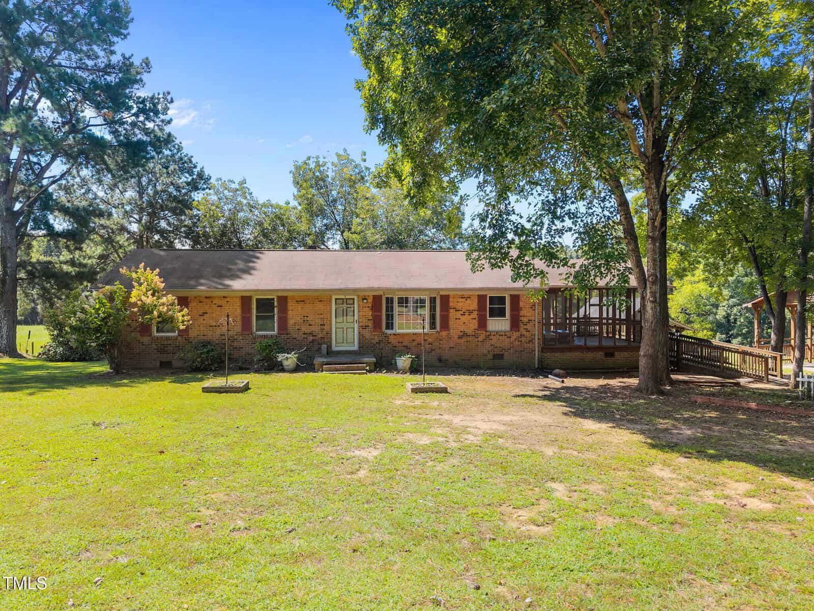 A single-story brick house with a covered front porch, green lawn, and several mature trees. The house has a gabled roof, white front door, and a wooden wheelchair-accessible ramp on the right side.