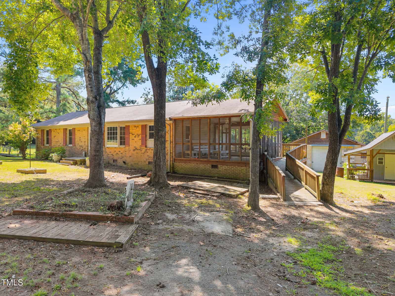 Single-story brick house with a screened porch and wooden ramp, surrounded by mature trees on a grassy lot. Sunlight filters through the leaves, casting shadows on the ground and the house exterior.