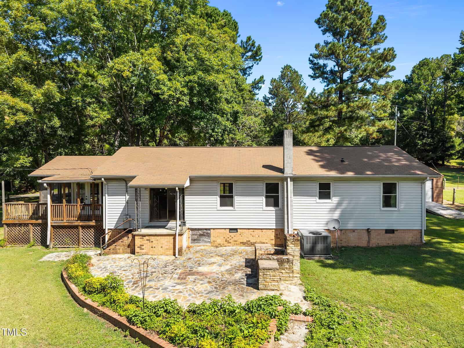 Single-story house with tan roof and white siding, elevated porch, stone patio, brick foundation, and surrounded by green grass, plants, and tall trees on a sunny day.