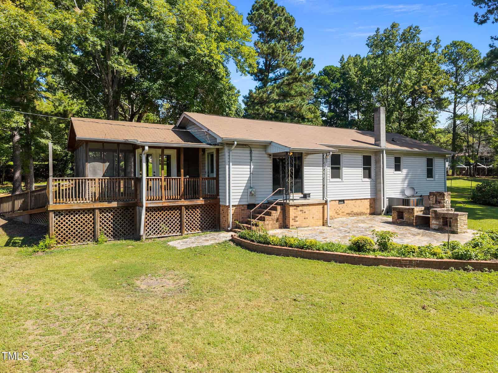 Single-story house with gray siding, brown roof, and a large wooden porch. There’s a stone patio with a built-in fire pit and seating, surrounded by green grass, trees, and a landscaped garden. Bright, sunny day.