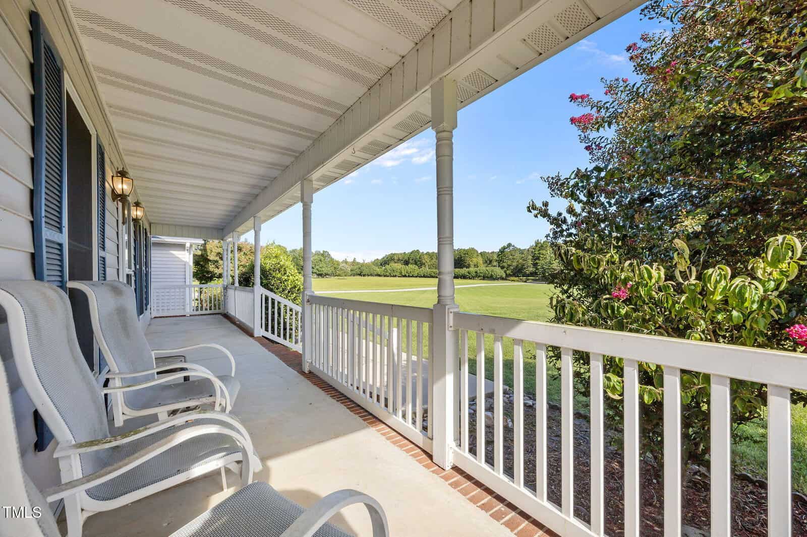 A covered front porch with white railings and several white rocking chairs overlooks a green lawn and trees under a clear blue sky. The porch ceiling features ventilation panels and hanging light fixtures.