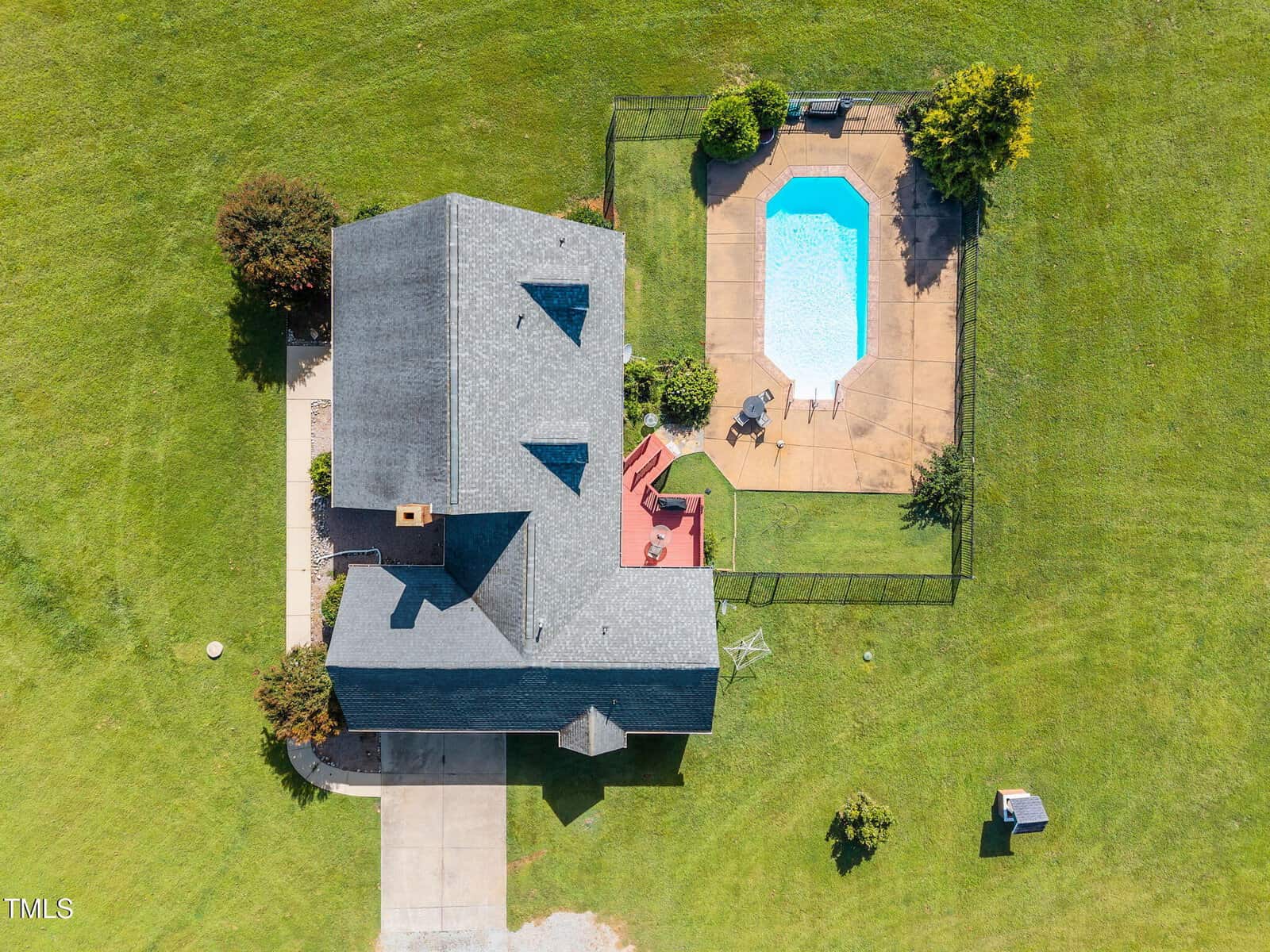 Aerial view of a house with a gray roof, a driveway, and a fenced-in backyard featuring a rectangular swimming pool, concrete patio, and grassy lawn. There are outdoor chairs and a small shed visible.