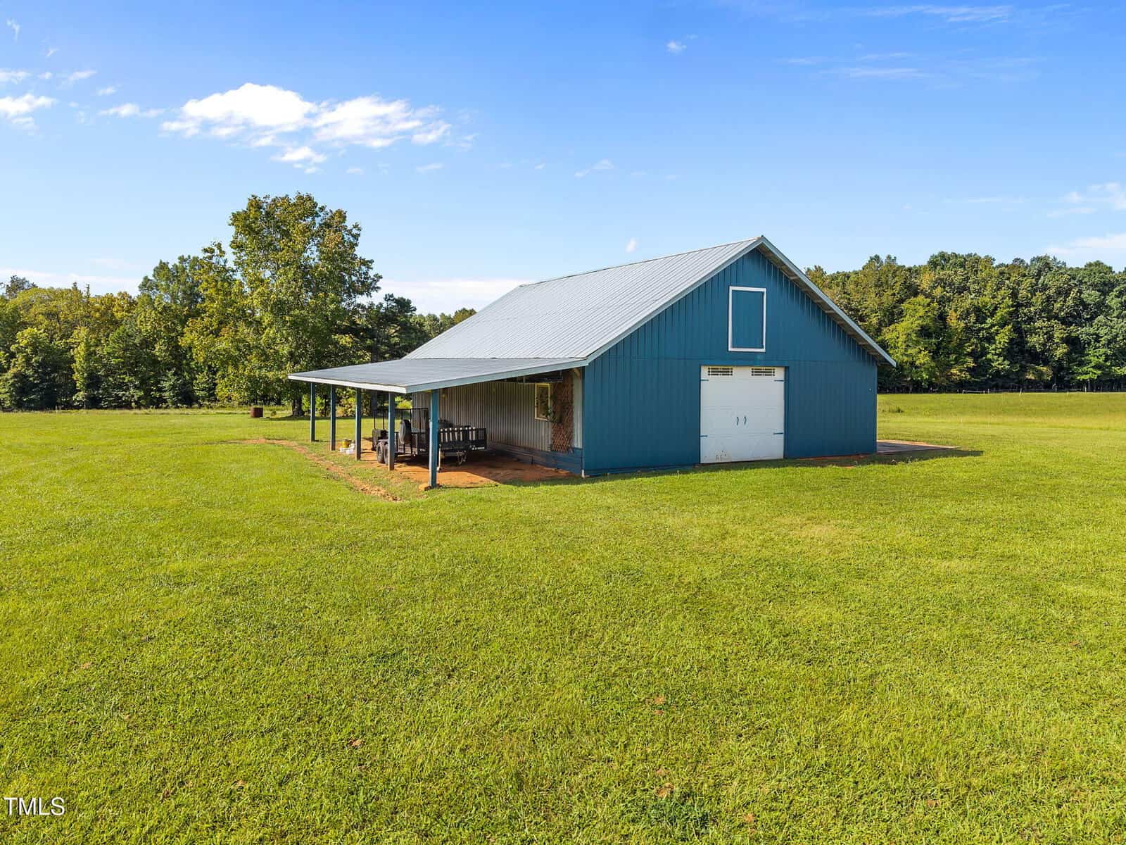 A blue metal barn with a gray roof sits on a large, open grassy field under a clear sky, with trees in the background and several chairs arranged under an overhang on one side of the building.