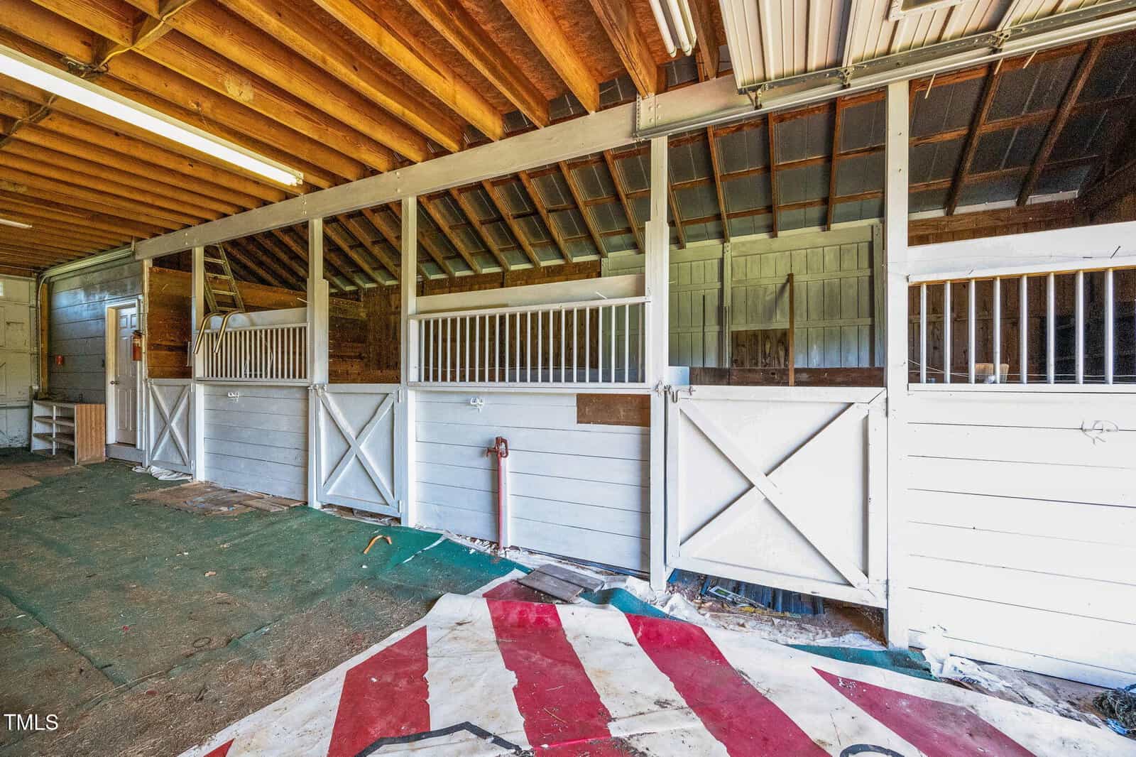 A view of an indoor horse stable with three white wooden stalls, each with a half-door and vertical bars. The ceiling has exposed wooden beams, and the floor is partially covered with old carpets and tarps.