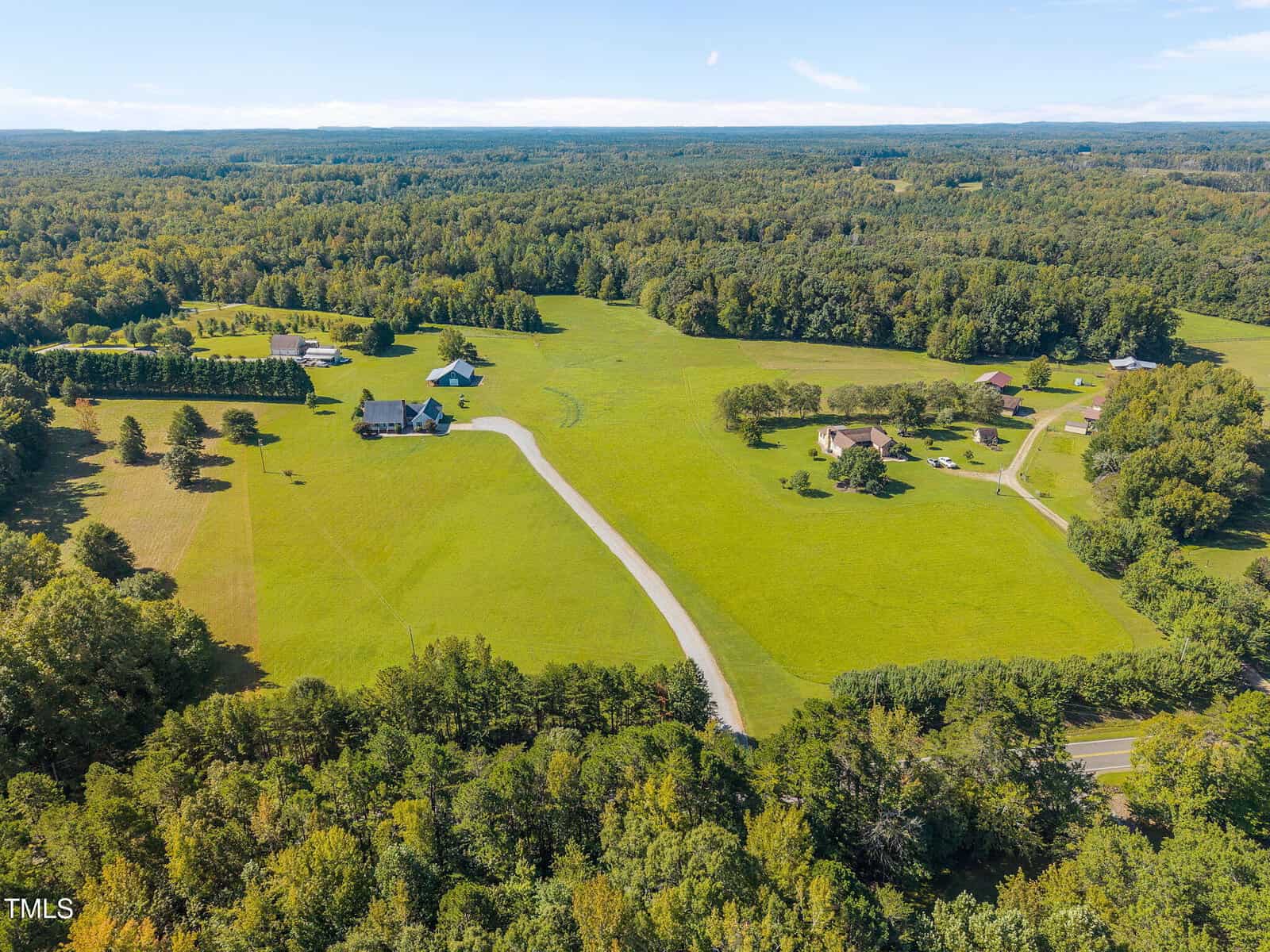 Aerial view of a rural landscape with several houses, green fields, trees, and a curved road running through the area; forested areas surround the open land.