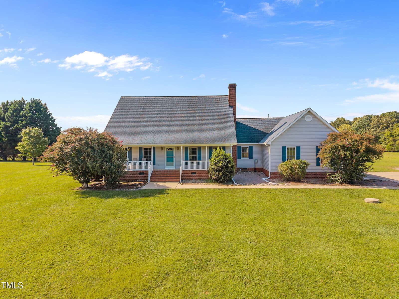 A single-story house with a large front porch, brick foundation, gable roof, and white siding sits on a spacious, well-maintained lawn with a few trees and shrubs under a blue sky.