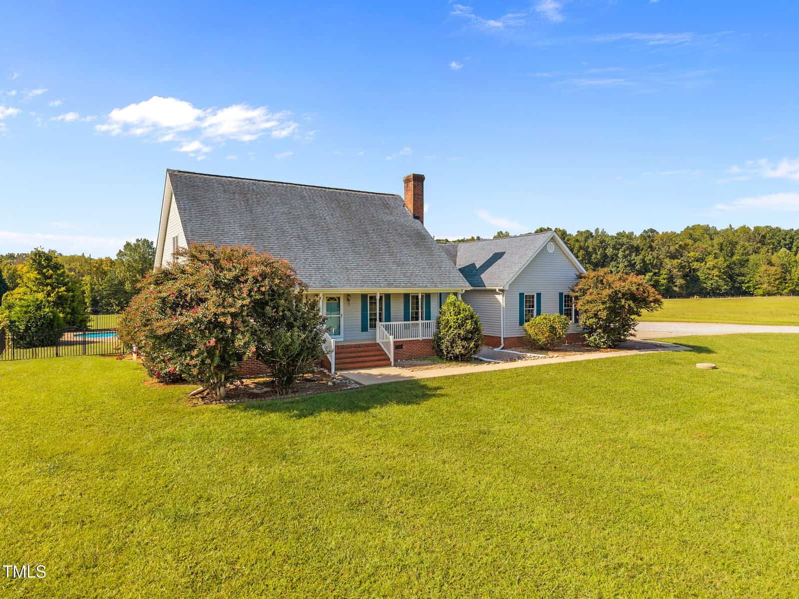A single-story house with a gray roof, brick chimney, and blue siding sits on a large, well-maintained grassy lawn with shrubs and trees in the front, under a clear blue sky.