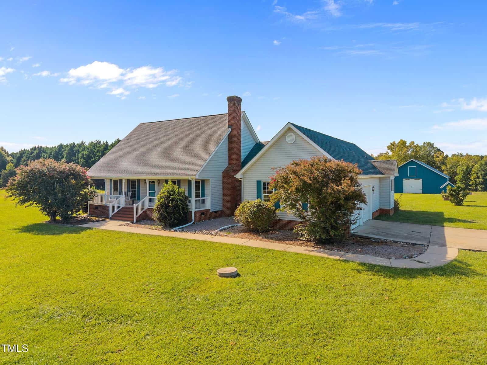 A single-story house with a large front porch, brick chimney, and attached garage is surrounded by a green lawn. A blue barn-like structure is visible in the background under a clear sky.