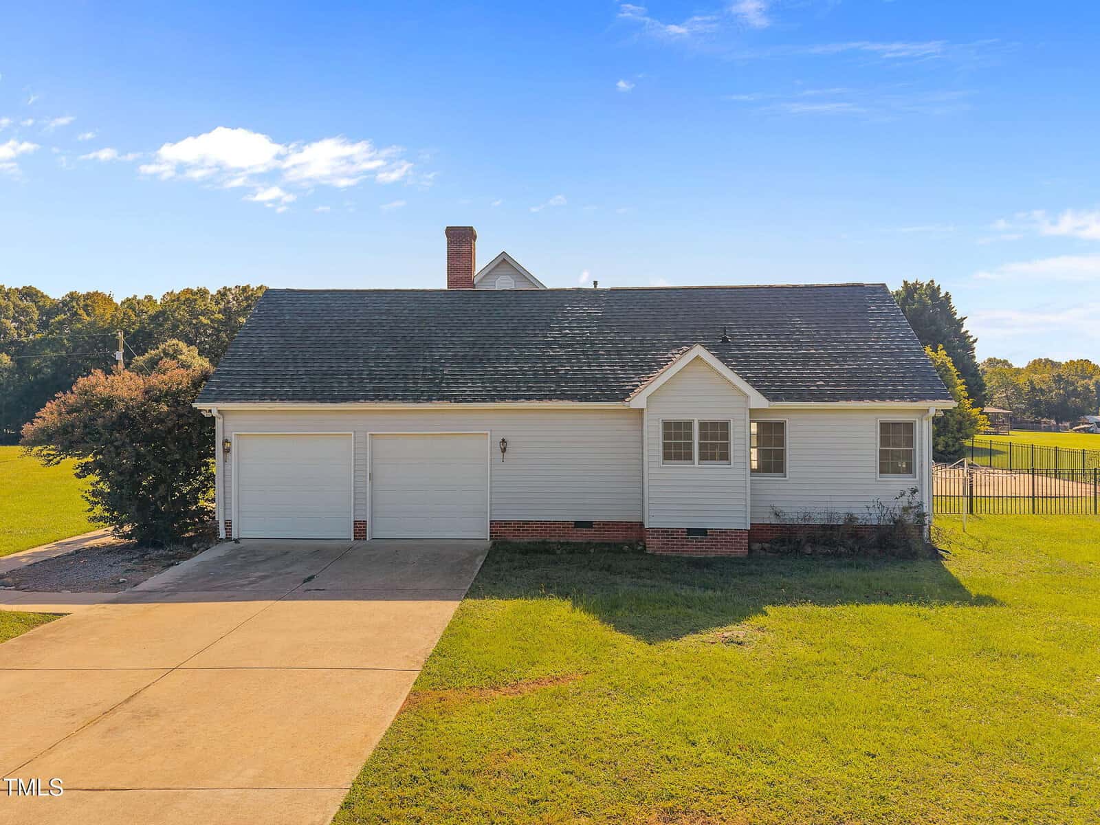 A single-story house with white siding, a dark roof, two-car garage, and brick accents sits on a green lawn under a clear blue sky. A concrete driveway leads to the garage. Trees and a fence are visible in the background.