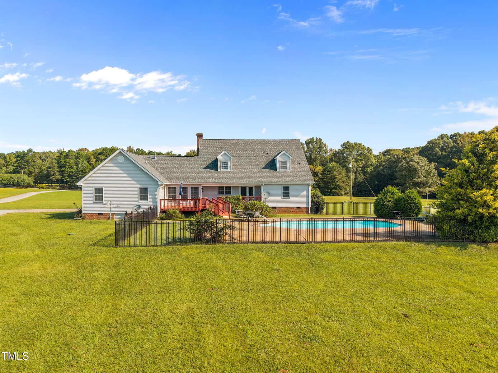 A white house with a gray roof sits on a large grassy lawn, featuring a fenced backyard with a swimming pool and surrounding trees under a blue sky with scattered clouds.