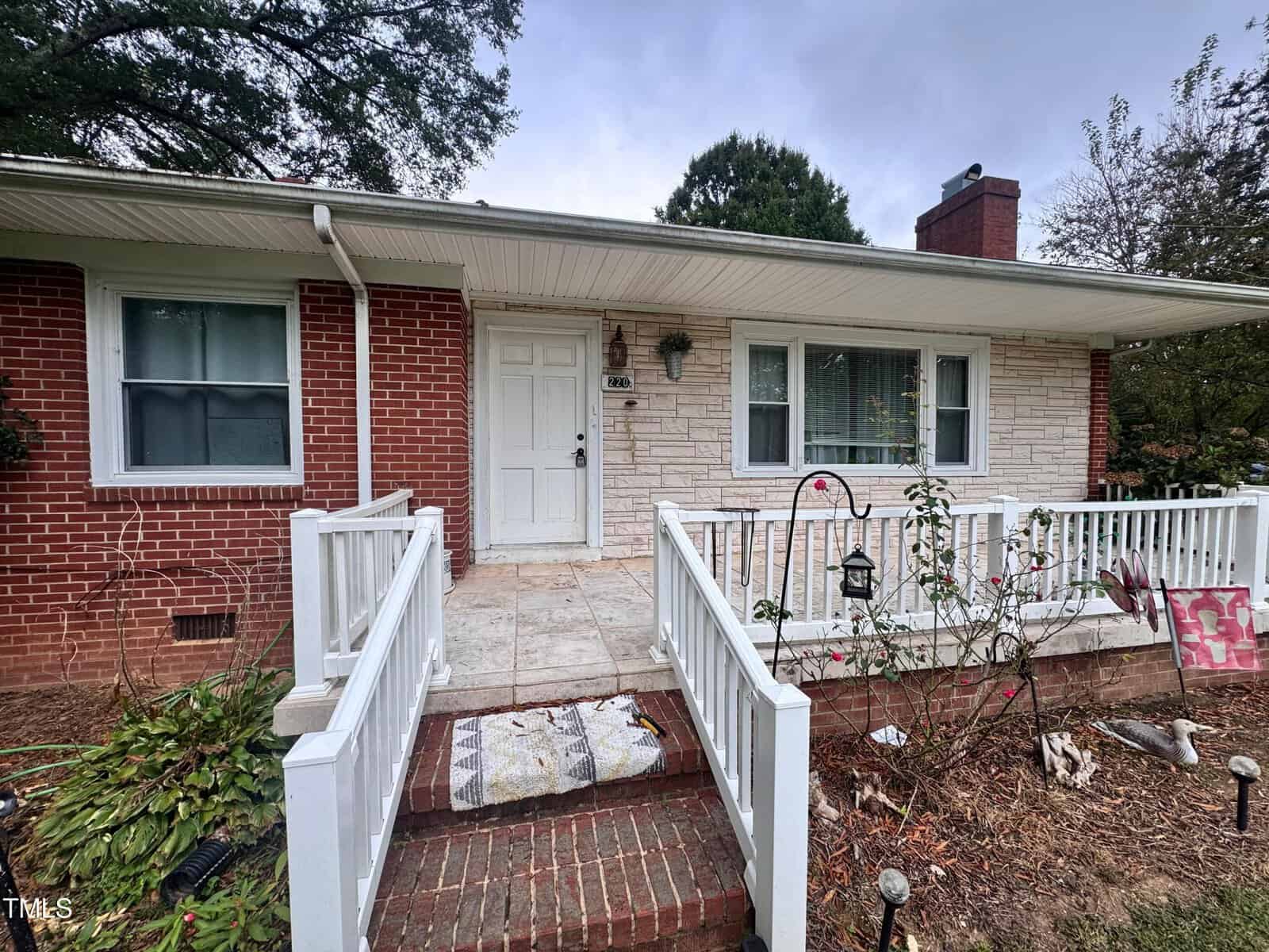 A single-story brick and stone house with a white front door, large front windows, and a white railing along a small porch. The front yard has scattered plants and a few items, with a paved walkway leading to the entrance.