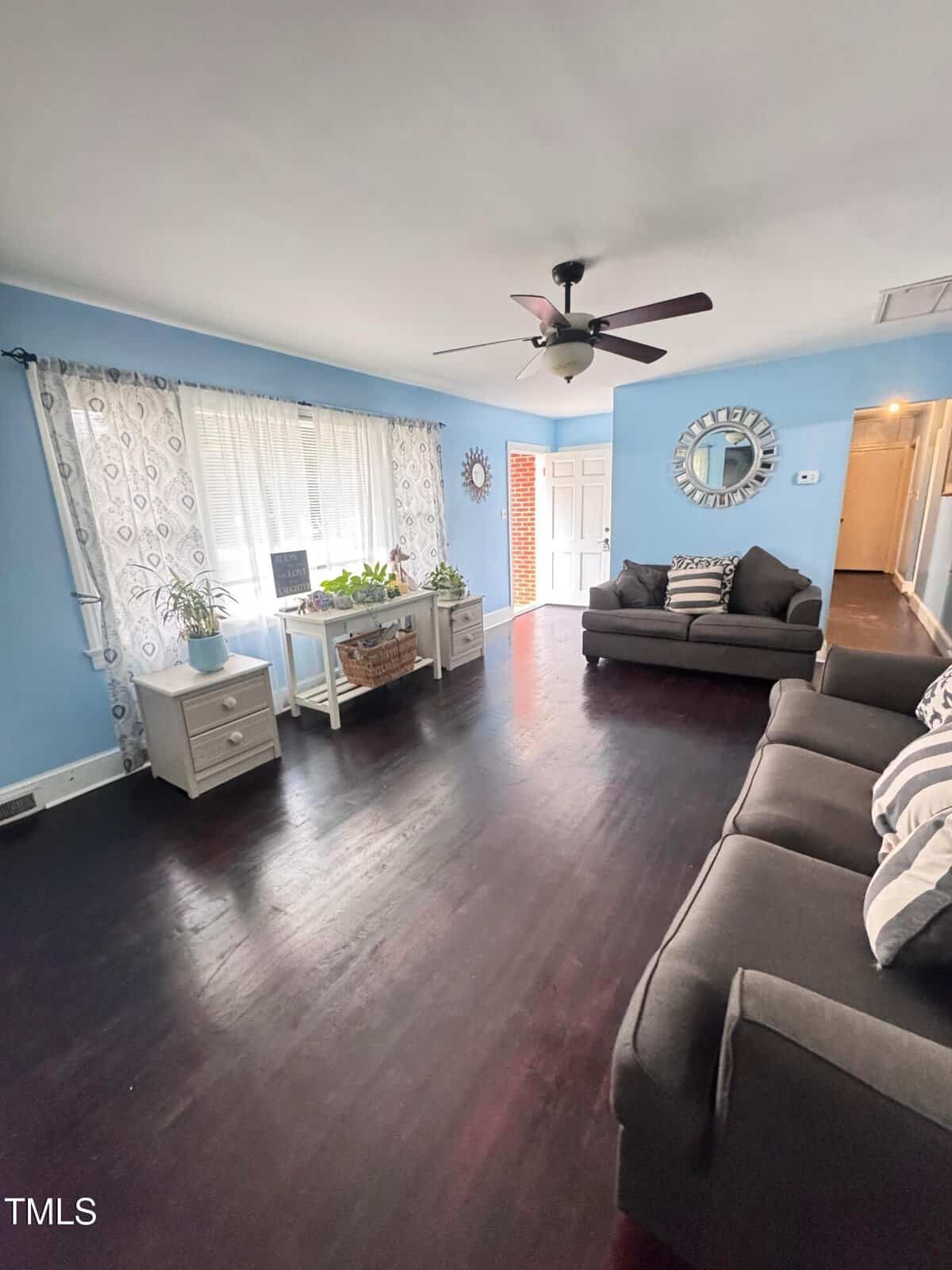 A living room with light blue walls, dark wood flooring, two gray sofas, white side tables, a ceiling fan, large window with sheer curtains, a wall mirror, and several potted plants near the window.