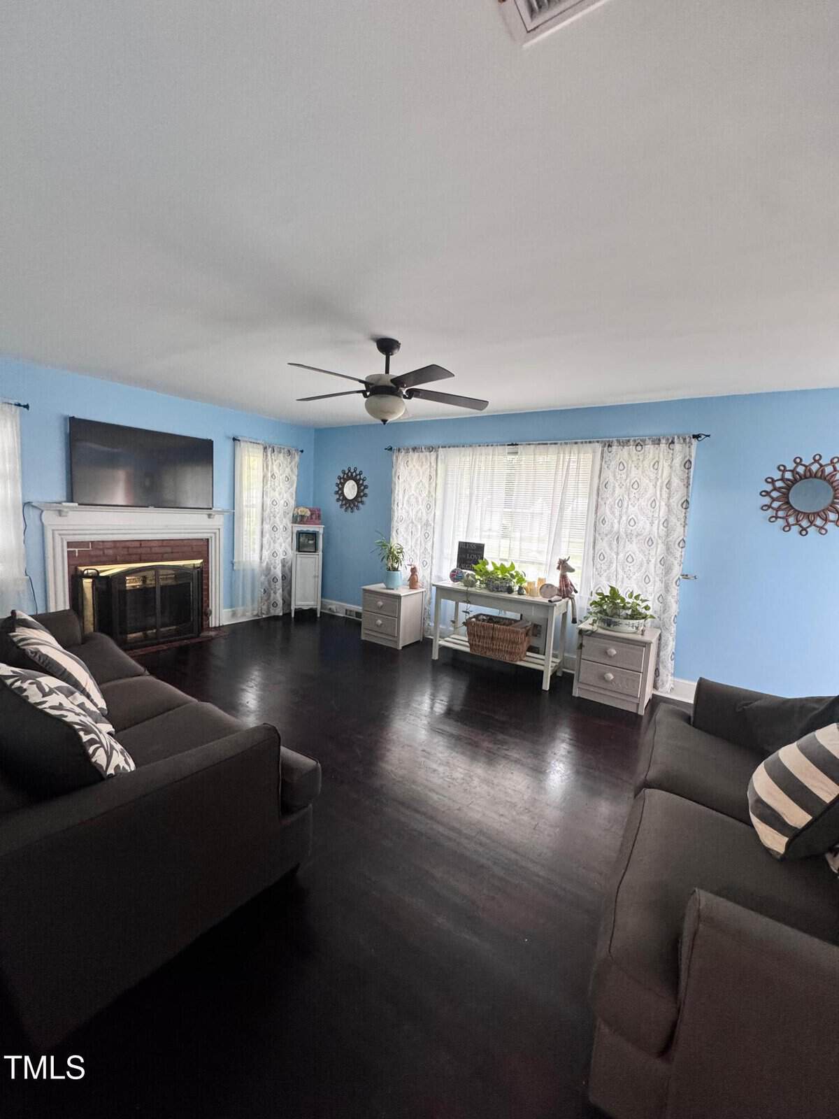 A living room with light blue walls, dark wood floors, two dark sofas, a fireplace with a TV above it, a ceiling fan, and a window with two desks underneath holding houseplants.