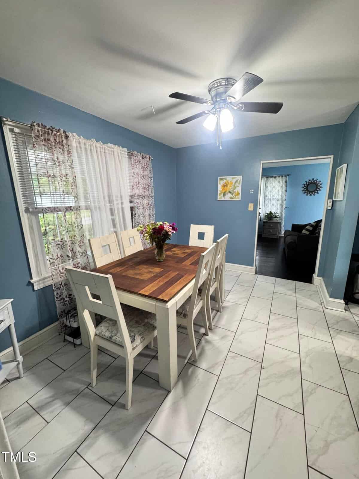 Dining room with blue walls, white tile floor, wooden table with six chairs, a vase of flowers, ceiling fan, and large window with patterned curtains. Doorway leads to another room with a dark sofa.