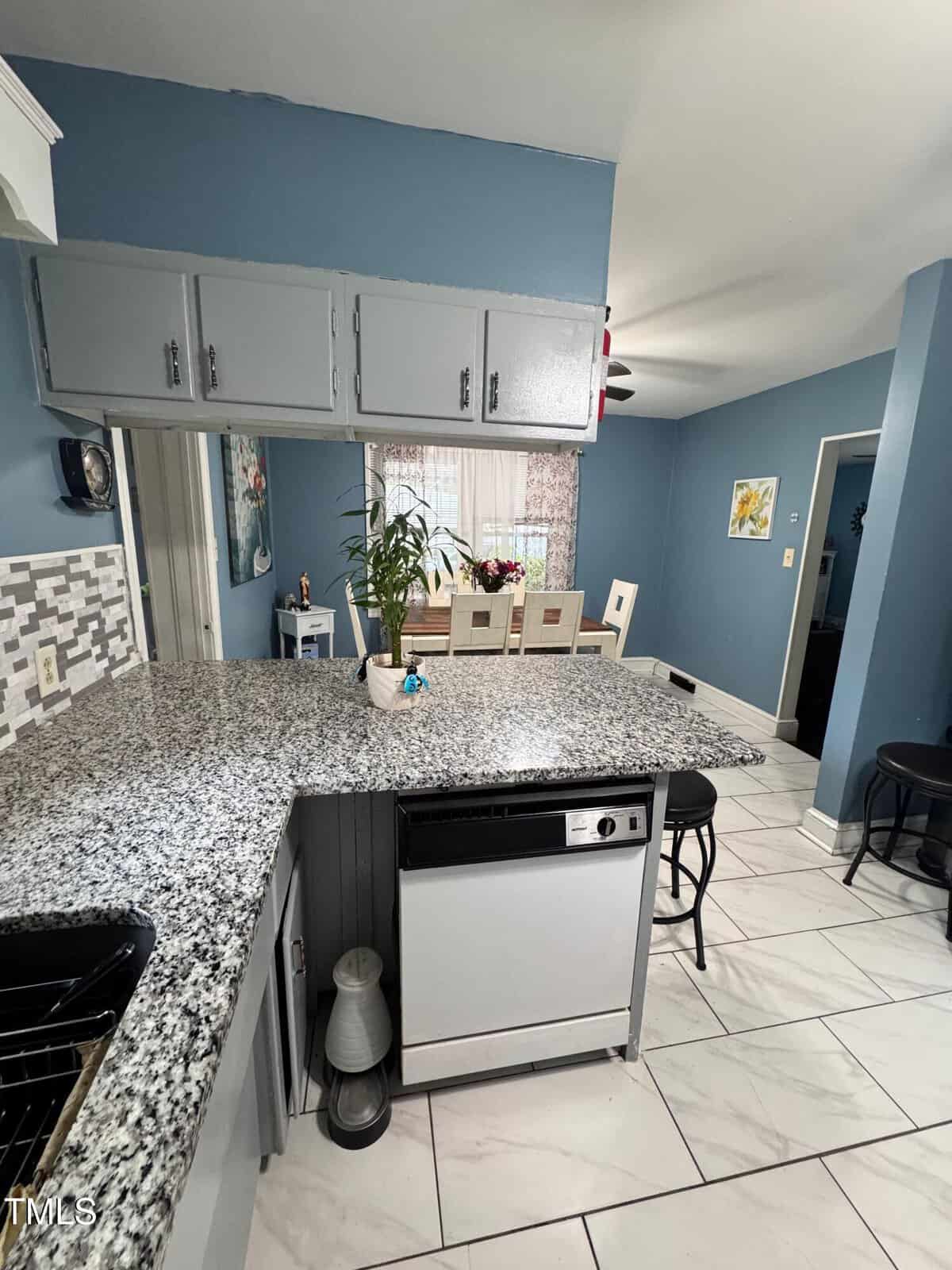 Bright kitchen with granite countertops, light gray cabinets, tile floor, and a small dining area with a white table and chairs. A plant and flowers decorate the space, and natural light streams through window curtains.