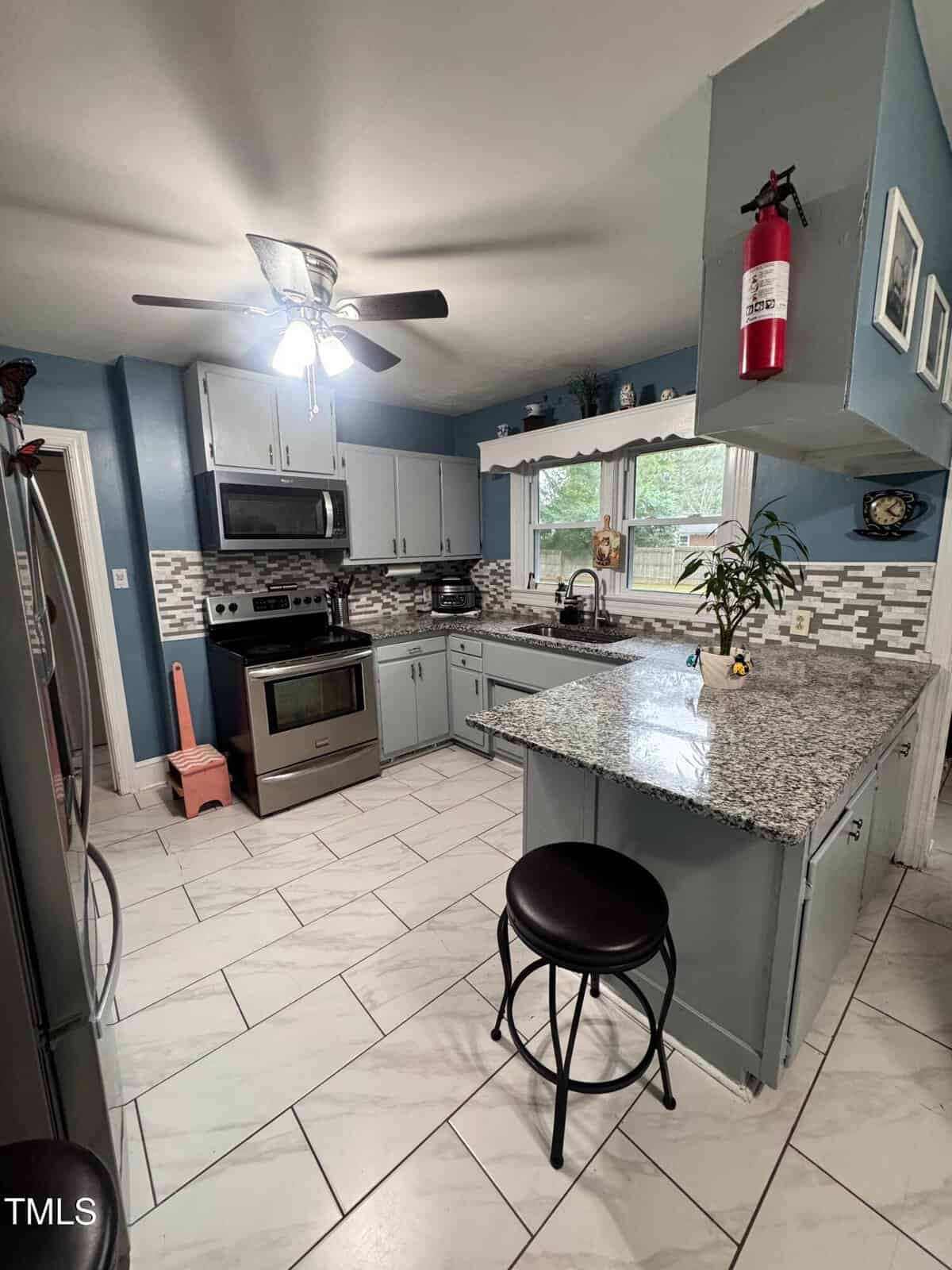 Modern kitchen with gray cabinets, stainless steel appliances, a barstool at a granite counter, a fire extinguisher on the wall, large window above the sink, and a pink broom near the door.