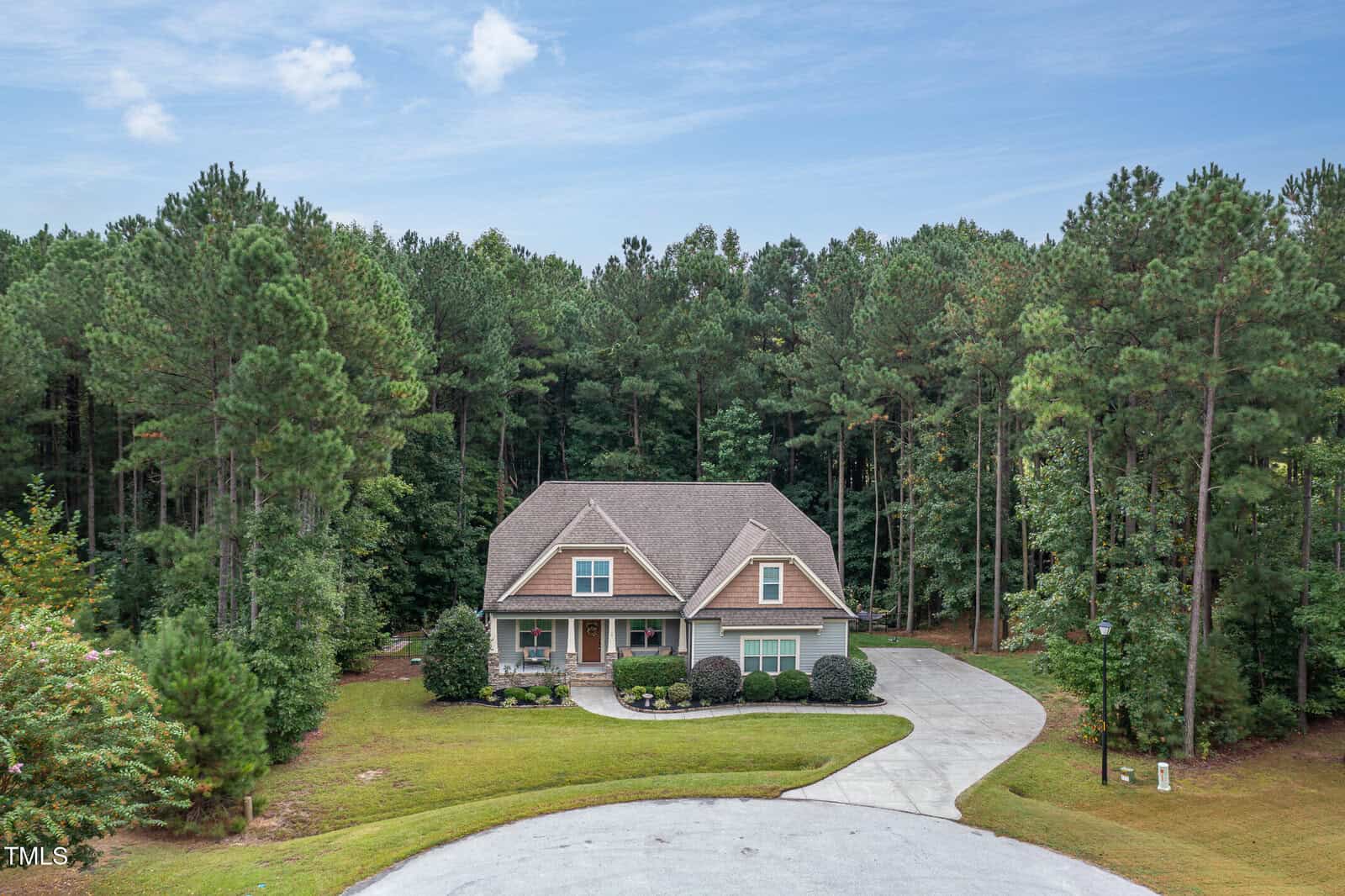A two-story house with a front porch and attached garage sits at the end of a cul-de-sac, surrounded by tall pine trees and greenery under a partly cloudy sky.
