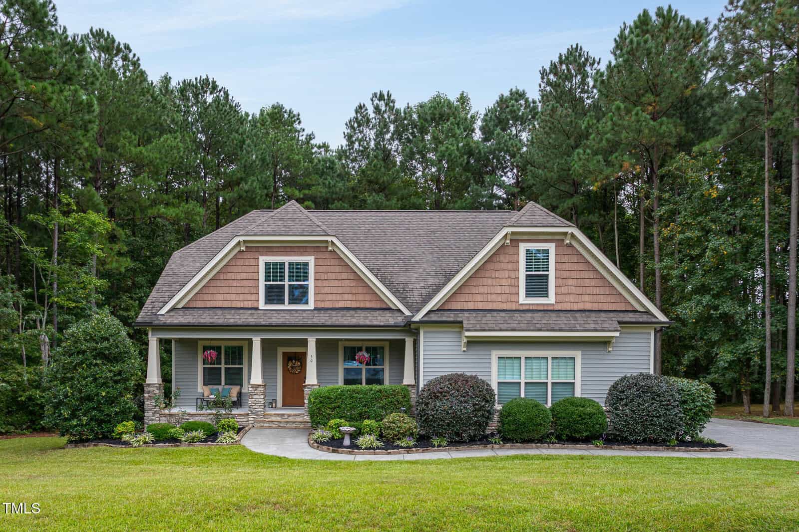 A single-family house with gray siding, brown shingles, and a covered front porch, surrounded by neatly trimmed bushes and trees, with a curved driveway and a wooded background.