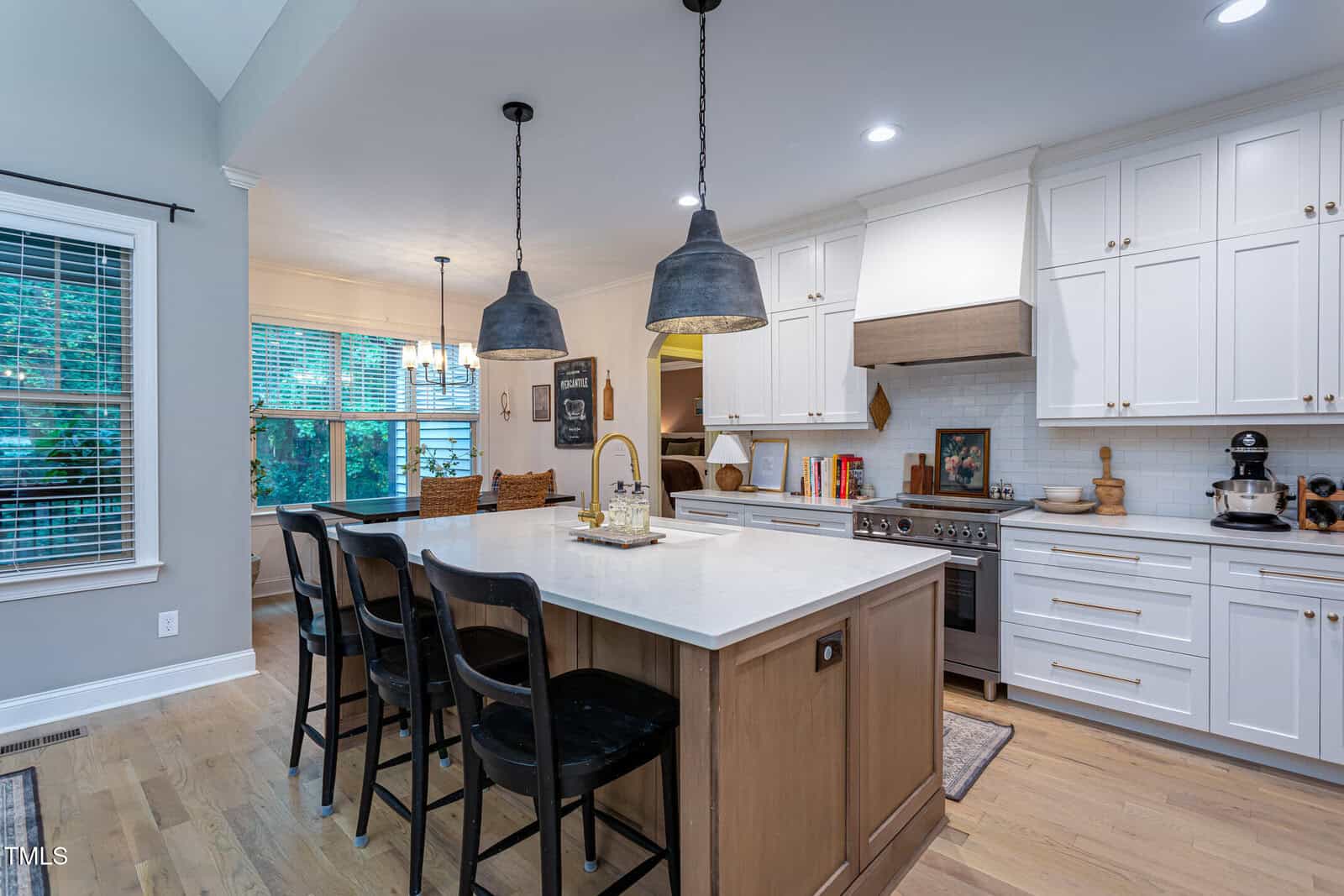 Modern kitchen with white cabinets, a large island with wood paneling, three black chairs, pendant lights, stainless steel appliances, and hardwood floors. Dining area with a table and windows is visible in the background.