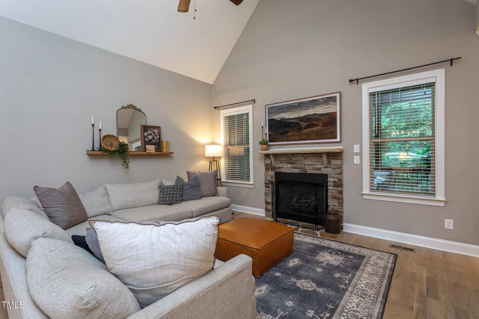 A living room with a light gray sectional sofa, a brown ottoman, a stone fireplace, a landscape painting above the mantel, shelves with decor, a navy rug, wood floors, and two windows with blinds letting in natural light.