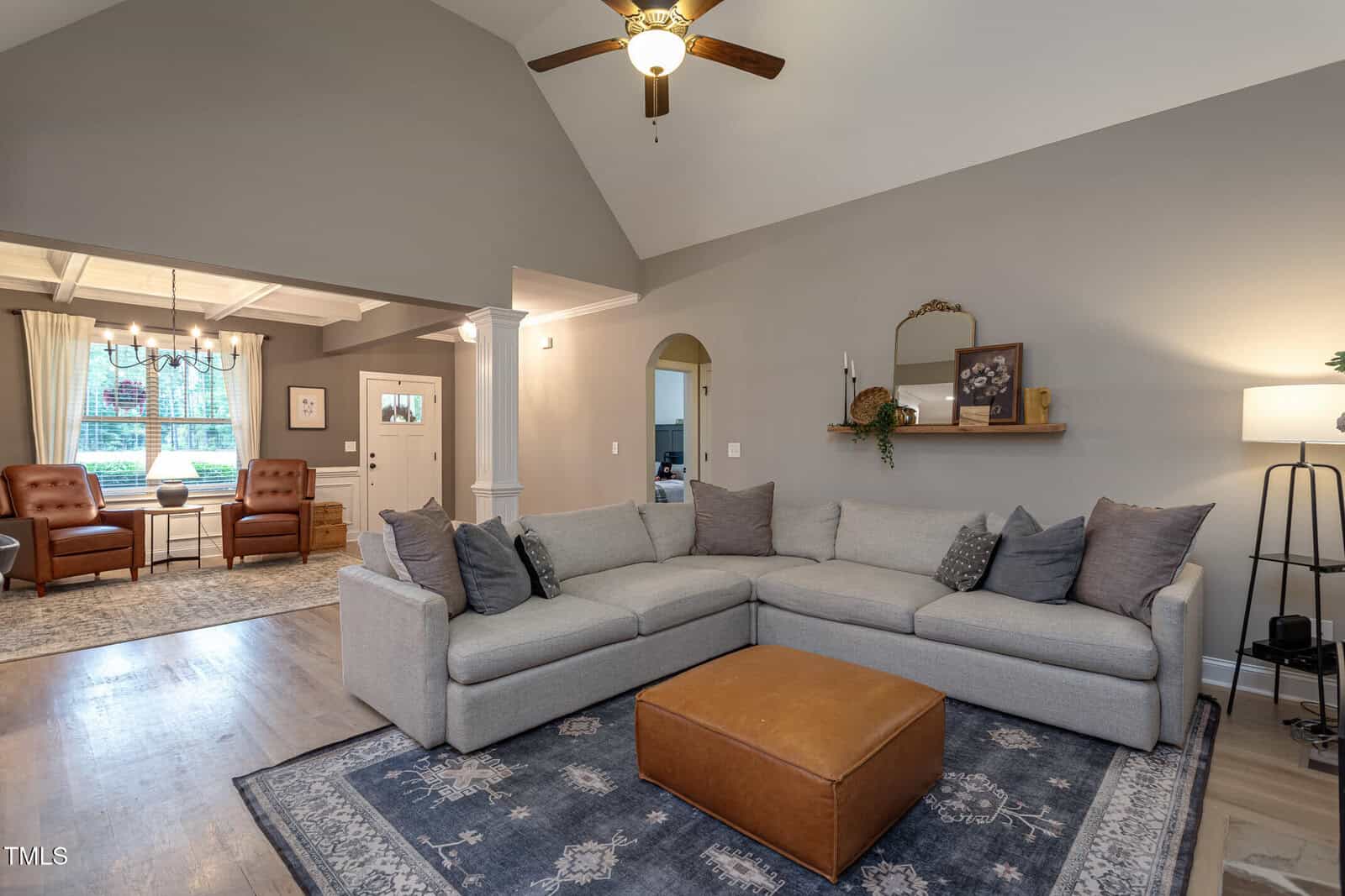 A living room with a gray sectional sofa, brown ottoman, and floor lamp on a patterned rug. Adjacent dining area features a table, chairs, and chandelier. Neutral wall colors and hardwood floors. Ceiling fan overhead.