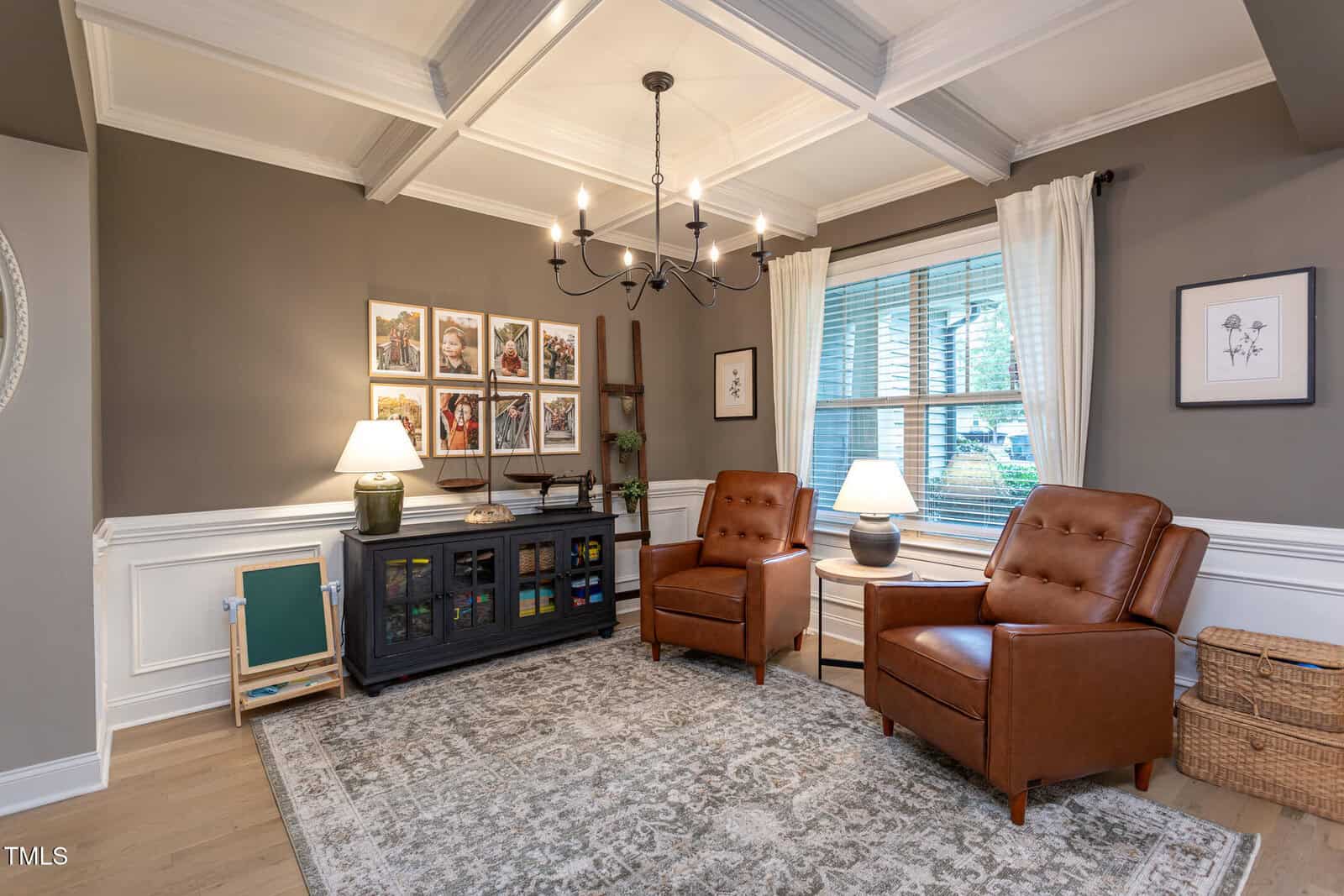 A cozy sitting room with two brown leather armchairs, a large rug, a dark cabinet with lamps, framed photos on the wall, a large window with blinds, and a black chandelier hanging from a coffered ceiling.
