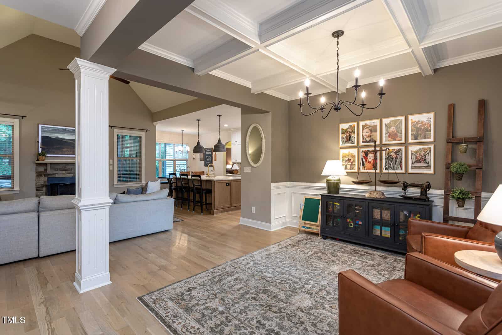 A spacious open-concept living area with a cozy sitting room, brown leather chairs, a patterned rug, wall art, and a dining area leading to a kitchen with pendant lights and wooden cabinets in the background.
